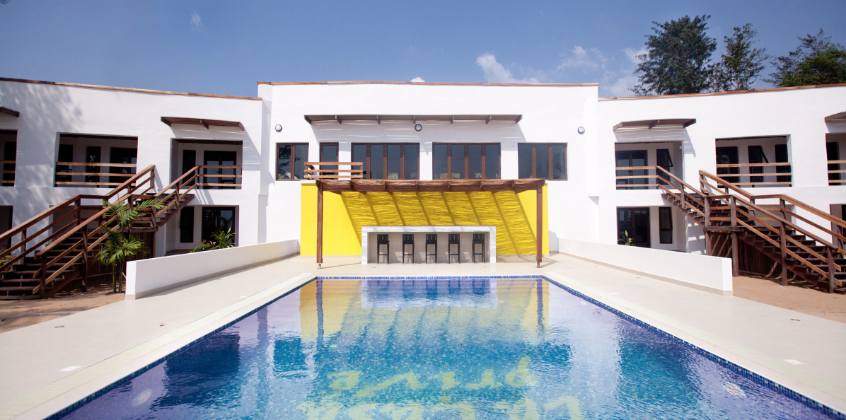 A sunny day at a tranquil holiday resort designed by Oshinowo Studio, the leading architectural design studio in Lagos, with a large swimming pool in the foreground and a white building with yellow accents,