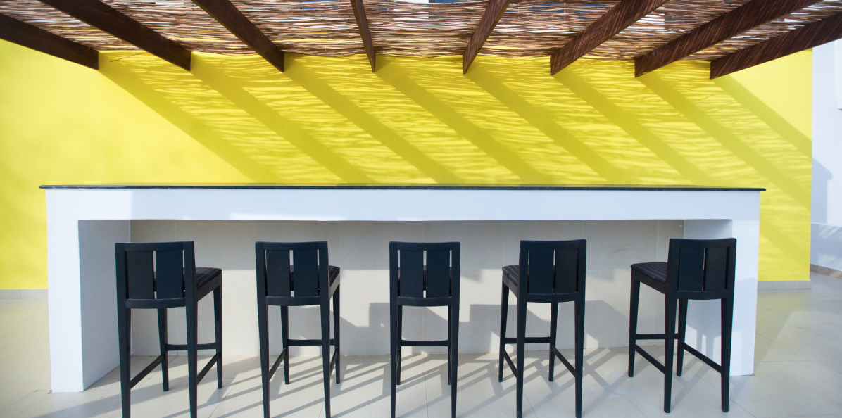 Modern minimalist bar setting with sleek black stools against a vibrant yellow wall, highlighted by an interesting shadow pattern from a unique wooden ceiling overhead, designed by Oshinowo Studio, a leading architectural design studio