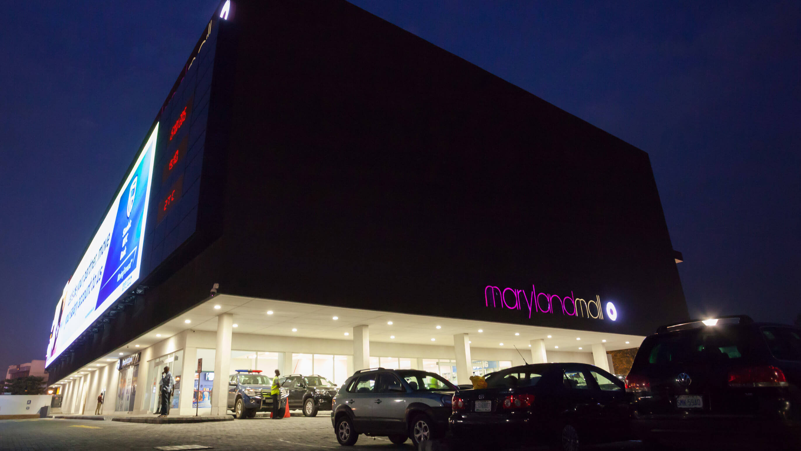 Evening view of Maryland Mall, designed by Oshinowo Studio and led by Tosin Oshinowo. The large, imposing structure of the mall is captured against the night sky, its facade dramatically illuminated by a brightly lit billboard showcasing advertisements, including a prominent one for Stanbic IBTC Bank. The mall's name is highlighted in pink neon letters on the side. Below, the entrance is brightly lit, with several cars parked in the foreground, adding a bustling atmosphere to the scene. The temperature display reads "27°C", suggesting a warm evening.