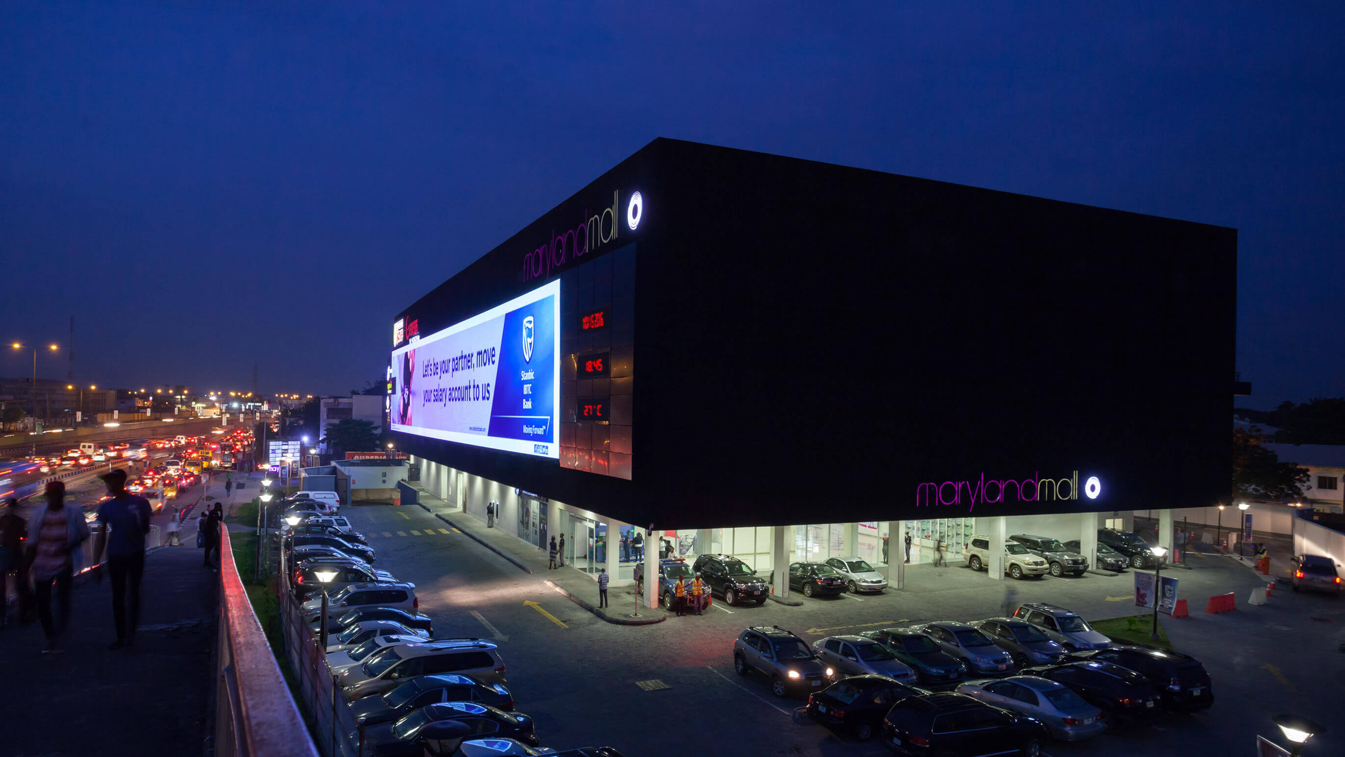 Nighttime view of Maryland Mall, designed by Oshinowo Studio and led by Tosin Oshinowo. The mall's exterior is striking with its large, illuminated billboard featuring an advertisement from Stanbic IBTC Bank. The facade is sleek and modern, primarily black, with the mall's name in stylish pink lettering. The parking lot in front of the mall is well-lit and filled with cars, showcasing a bustling evening scene. A digital display on the building provides the time "18:59" and temperature "27°C", adding a dynamic urban touch to the setting.
