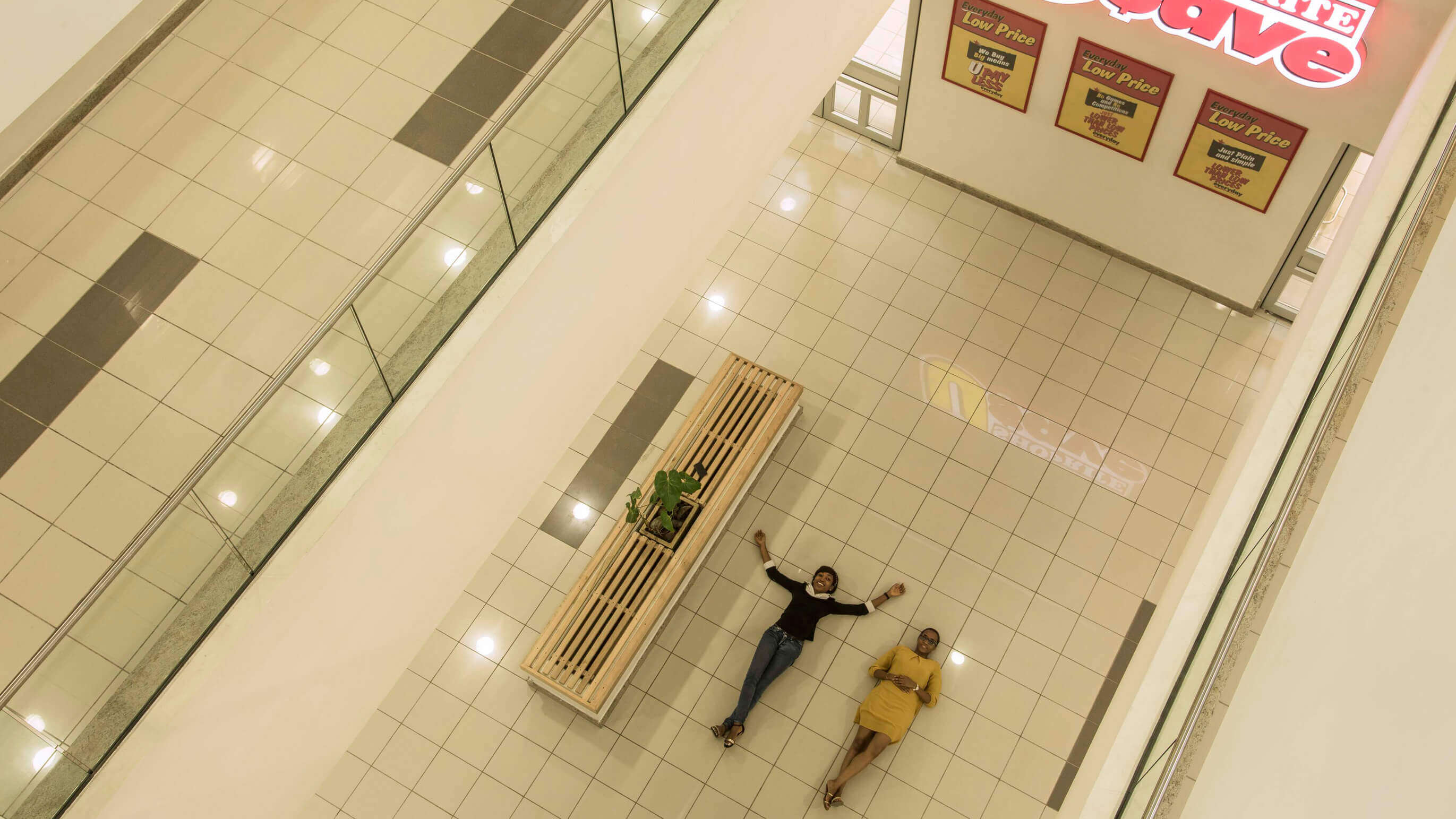 An overhead view of a spacious interior within a shopping mall designed by Oshinowo Studio and led by Tosin Oshinowo. Two individuals are playfully lying on the floor, their arms spread wide, next to a wooden bench with a small plant. Above them hangs a sign for Shoprite U-Save, promoting "Everyday Low Price." The floor is tiled in a light color with darker accents, reflecting the bright interior lighting. The area is bordered by a balustrade, providing a glimpse into another level of the mall, contributing to the open and airy architecture of the space.