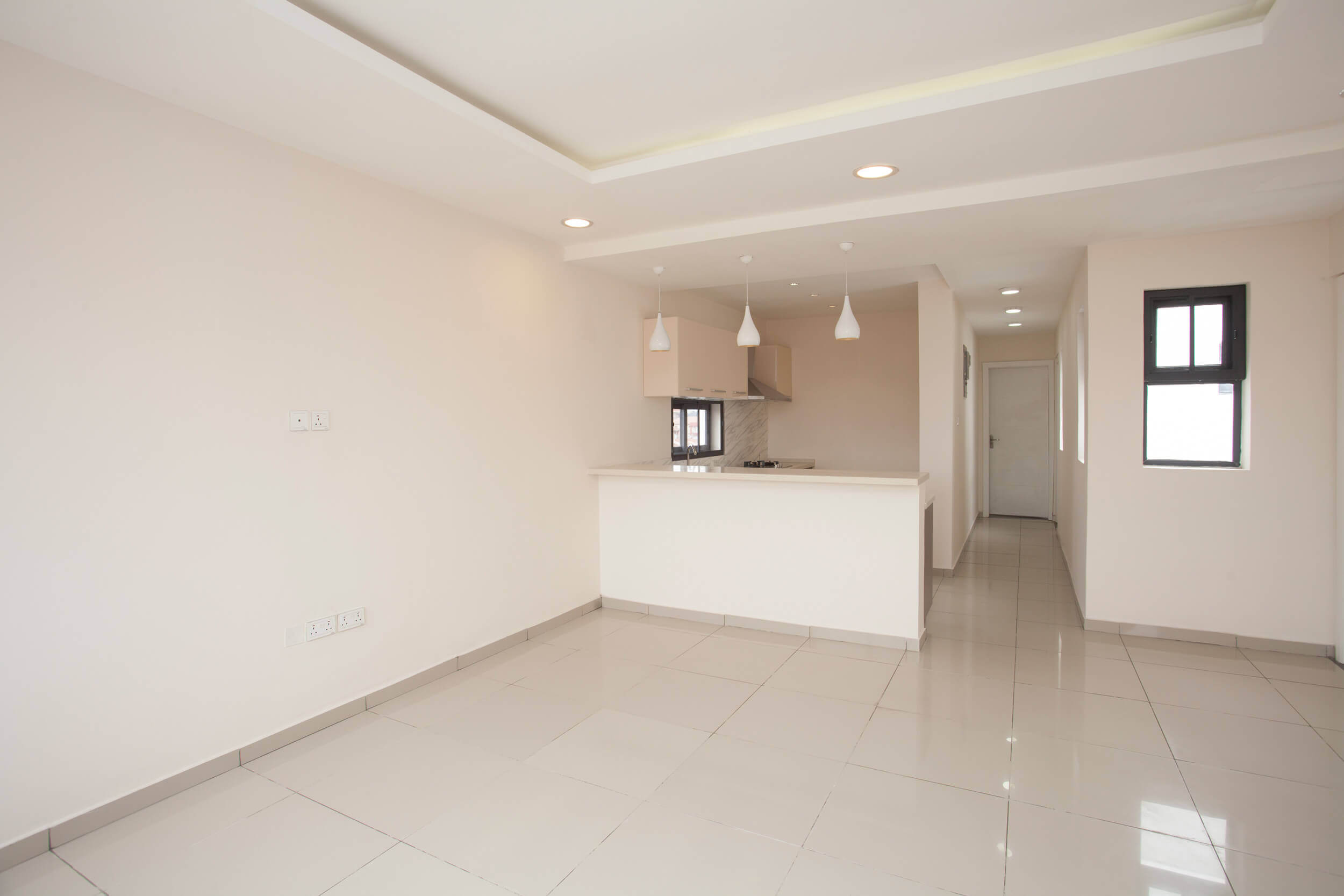 Bright and airy open-plan living space in The Plum Apartments Lagos, featuring a gleaming white tiled floor that complements the soft beige walls and white ceiling with recessed lighting. The kitchen area is visible in the background, defined by its clean lines, modern pendant lighting, and a breakfast bar with a marble-effect countertop that adds a touch of elegance. The space is designed for a seamless blend of comfort and style, offering a canvas for personalization and decor. Project led by Tosin Oshinowo