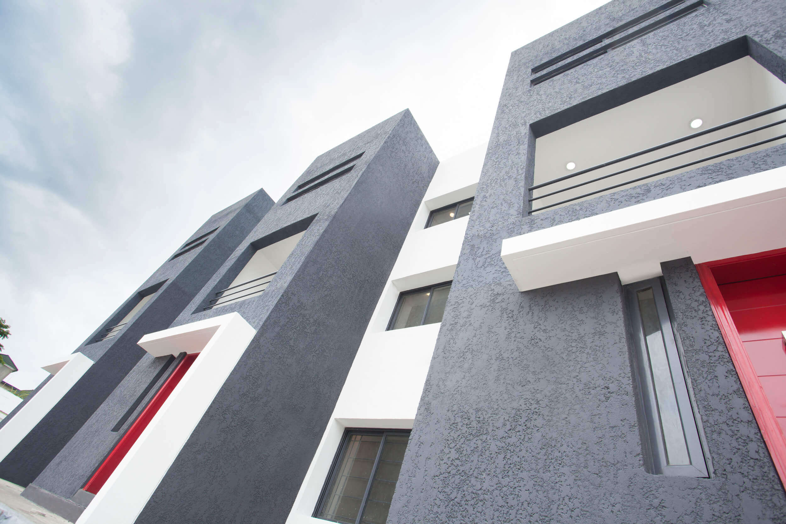 Dynamic exterior view of The Plum Apartments Lagos, showcasing a bold architectural design with textured dark gray façades contrasted by bright red doors, white trimmings, and modern balconies. The striking color palette and angular lines create a contemporary urban aesthetic, while a person in motion adds a vibrant human element to the scene. Project led by Tosin Oshinowo