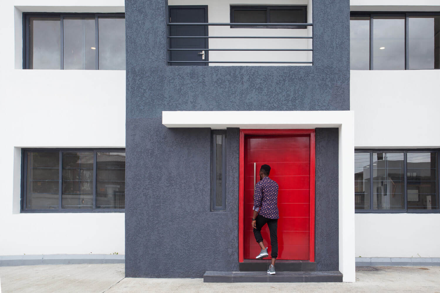 A person is captured in motion as they step through the vibrant red doorway of a modern building with a dark gray and white facade. The building features a balanced arrangement of large windows and geometric shapes, providing a contrast of colors and a contemporary look. Project led by Tosin Oshinowo