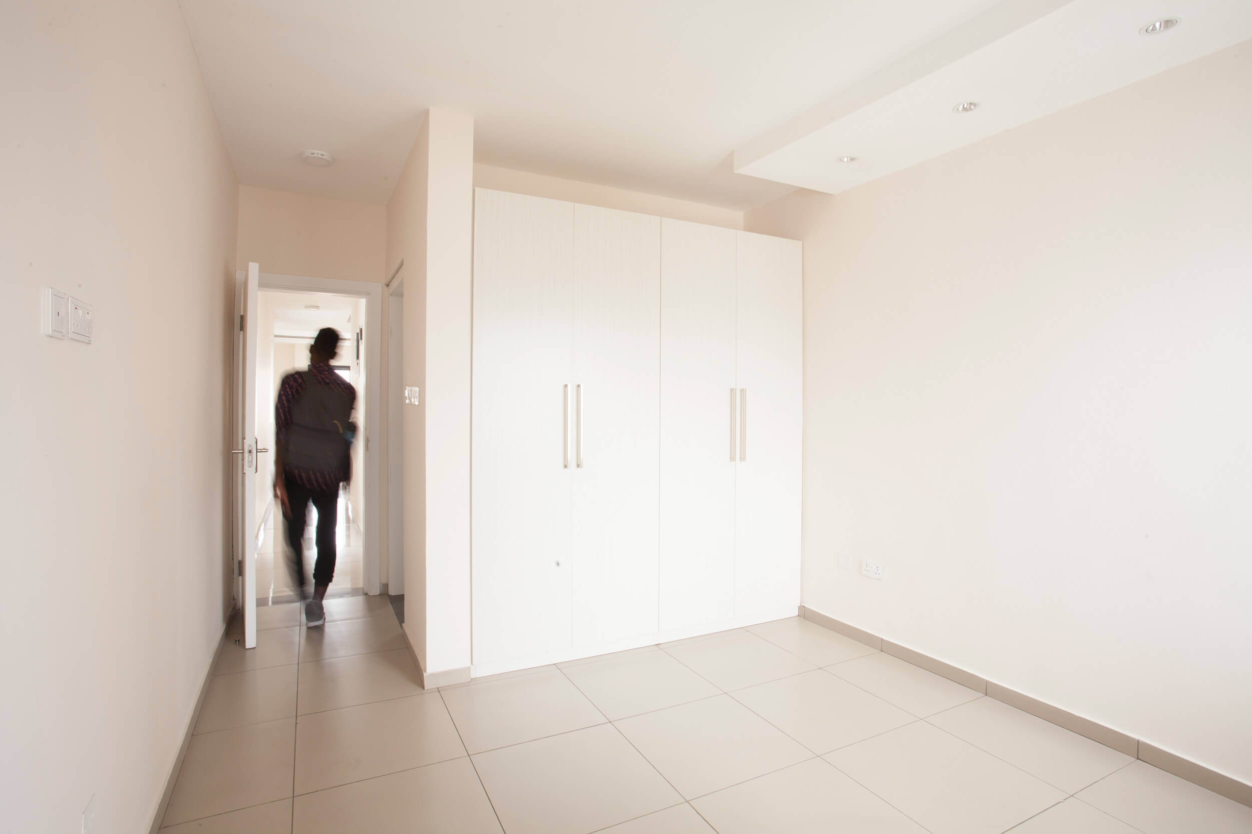 A minimalist bedroom within an apartment, featuring large white wardrobes with sleek silver handles that complement the room’s modern aesthetic. The space is well-lit, with recessed lighting in the ceiling adding to the clean lines and bright atmosphere. The tile flooring is a gentle cream, reflecting the light and enhancing the sense of spaciousness, while a figure is seen moving through the doorway, adding a sense of life and activity. Project led by Tosin Oshinowo