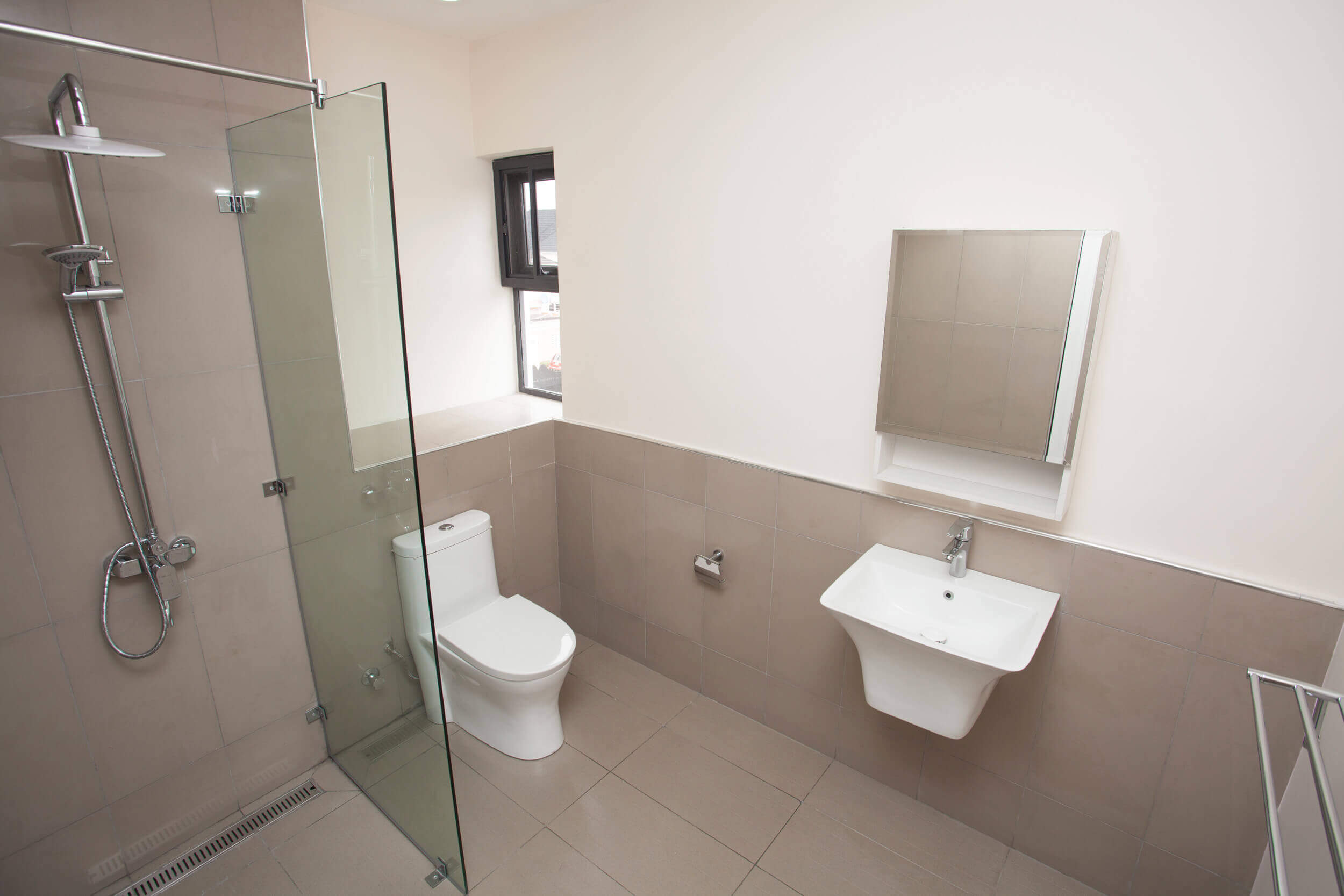 Modern and simplistic bathroom design with beige wall tiles and floor, featuring a walk-in shower with a clear glass partition, a white pedestal sink, and a classic toilet. The room is equipped with contemporary fixtures and a small window allowing for natural light and ventilation, creating a clean and bright space. Project led by Tosin Oshinowo