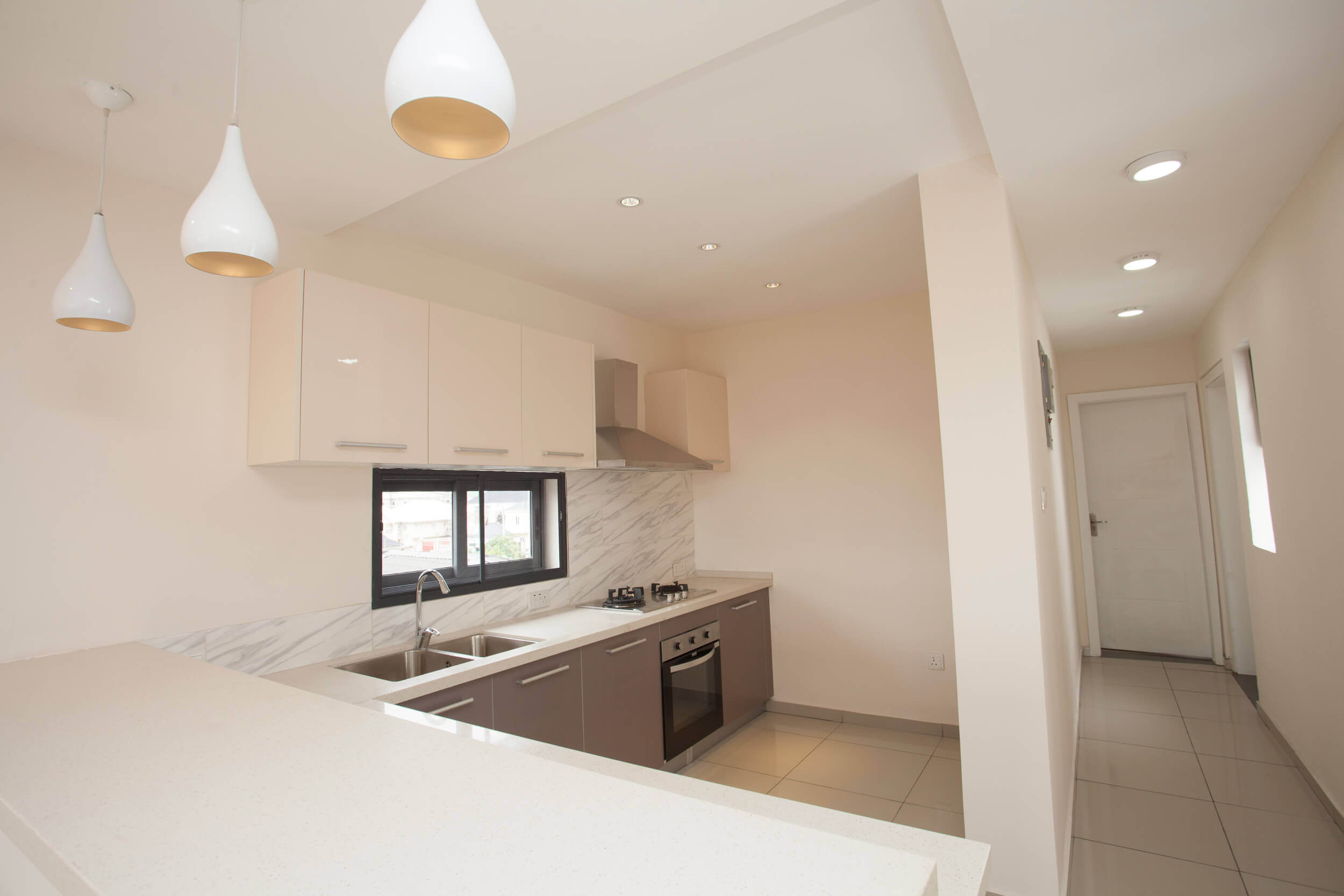 Elegant modern kitchen inside The Plum Apartments Lagos, featuring pale beige cabinetry and marble-like backsplash, complemented by a white quartz countertop. The space is softly illuminated by natural light and chic pendant lights with a golden interior, creating a warm, inviting atmosphere. The clean design lines and uncluttered space reflect a sophisticated urban living experience. Project led by Tosin Oshinowo