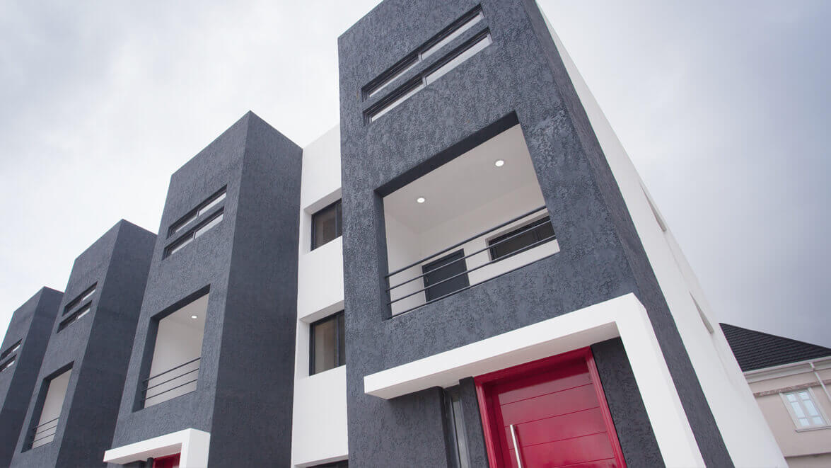 Dynamic exterior view of The Plum Apartments Lagos, showcasing a bold architectural design with textured dark gray façades contrasted by bright red doors, white trimmings, and modern balconies. The striking color palette and angular lines create a contemporary urban aesthetic, while a person in motion adds a vibrant human element to the scene. Project led by Tosin Oshinowo