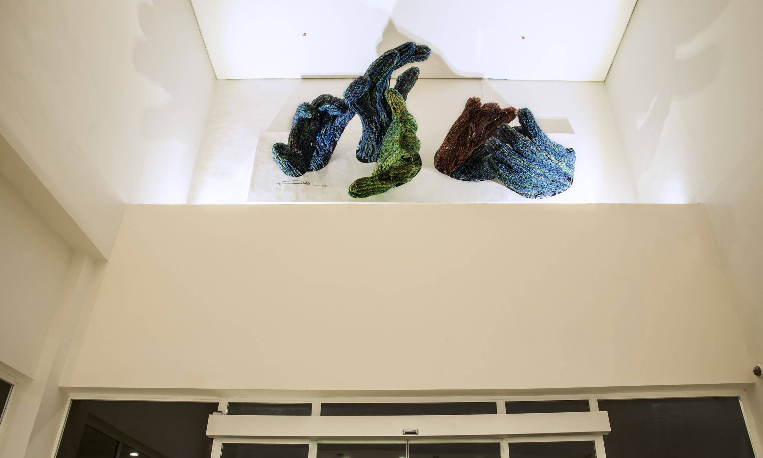 An artistic installation on the high ceiling of a building designed by Oshinowo Studio and led by Tosin Oshinowo. The artwork features several large, abstract sculptures made from what appears to be twisted and woven materials, in hues of blue, green, and brown. These dynamic forms are mounted against a stark white background, creating a striking visual contrast. The installation is viewed from below, emphasizing its position above a wide, uncluttered hallway within the modern architectural space, enhancing the aesthetic appeal of the building's interior design.