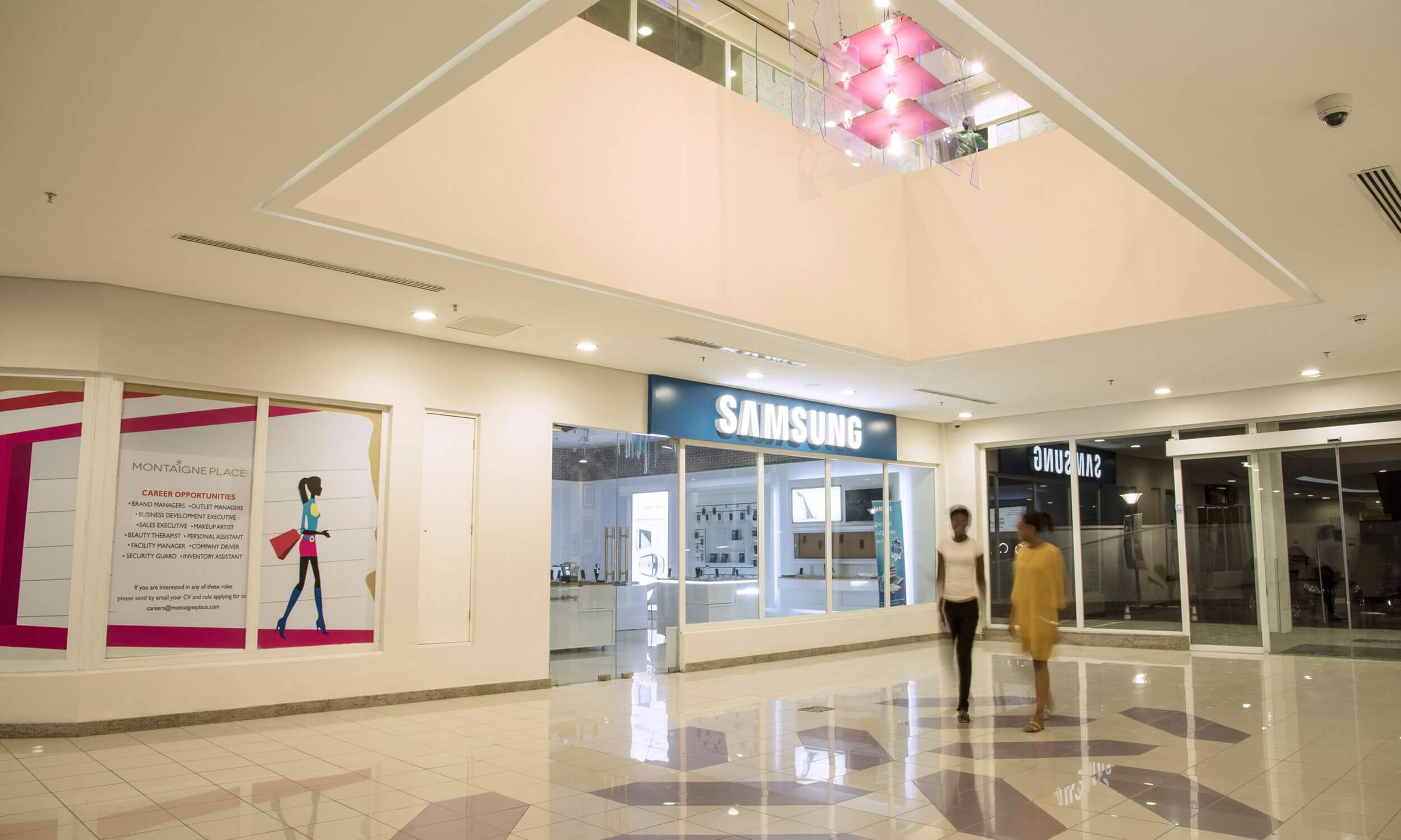 Inside Maryland Mall, designed by Oshinowo Studio and led by Tosin Oshinowo, this image captures a well-lit, spacious interior hallway with glossy tiled floors reflecting overhead lights. To the left, there's a shop with a colorful display promoting career opportunities at Montaigne Place. Straight ahead, the Samsung store showcases its sleek, modern interior through large glass windows. On the right, another store named "Samsung" is visible. The scene includes a few shoppers walking through, adding a dynamic element to the tranquil shopping environment. A unique pink light fixture hangs from the ceiling, adding a touch of modern design to the space.