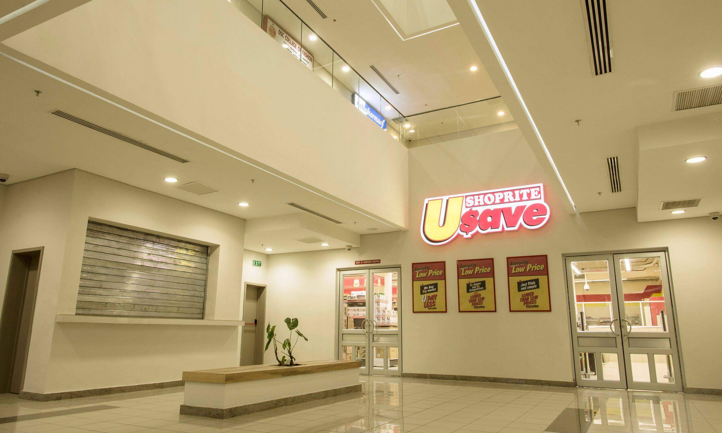 Inside a modern shopping mall designed by Oshinowo Studio and led by Tosin Oshinowo, this image showcases a spacious interior with a Shoprite U-Save store. The store features a bright red and yellow sign above its entrance, proclaiming "Everyday Low Price." The area is well-lit with recessed ceiling lights, and the floors are clean and shiny, reflecting the overhead lighting. A closed security window and a seating area with a small plant add to the utilitarian yet inviting atmosphere of the mall corridor.