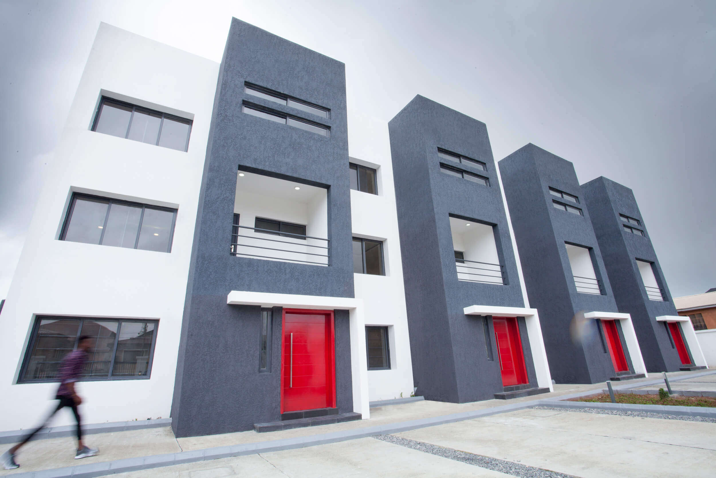 A contemporary and striking facade of The Plum Apartments Lagos, showcasing a modern architectural style with a combination of white and dark gray textures. The building features sharp geometric lines, large windows, and bold red doors that provide a pop of color against the muted tones. A blurred figure in motion adds a dynamic element to the scene, emphasizing the building's urban context.