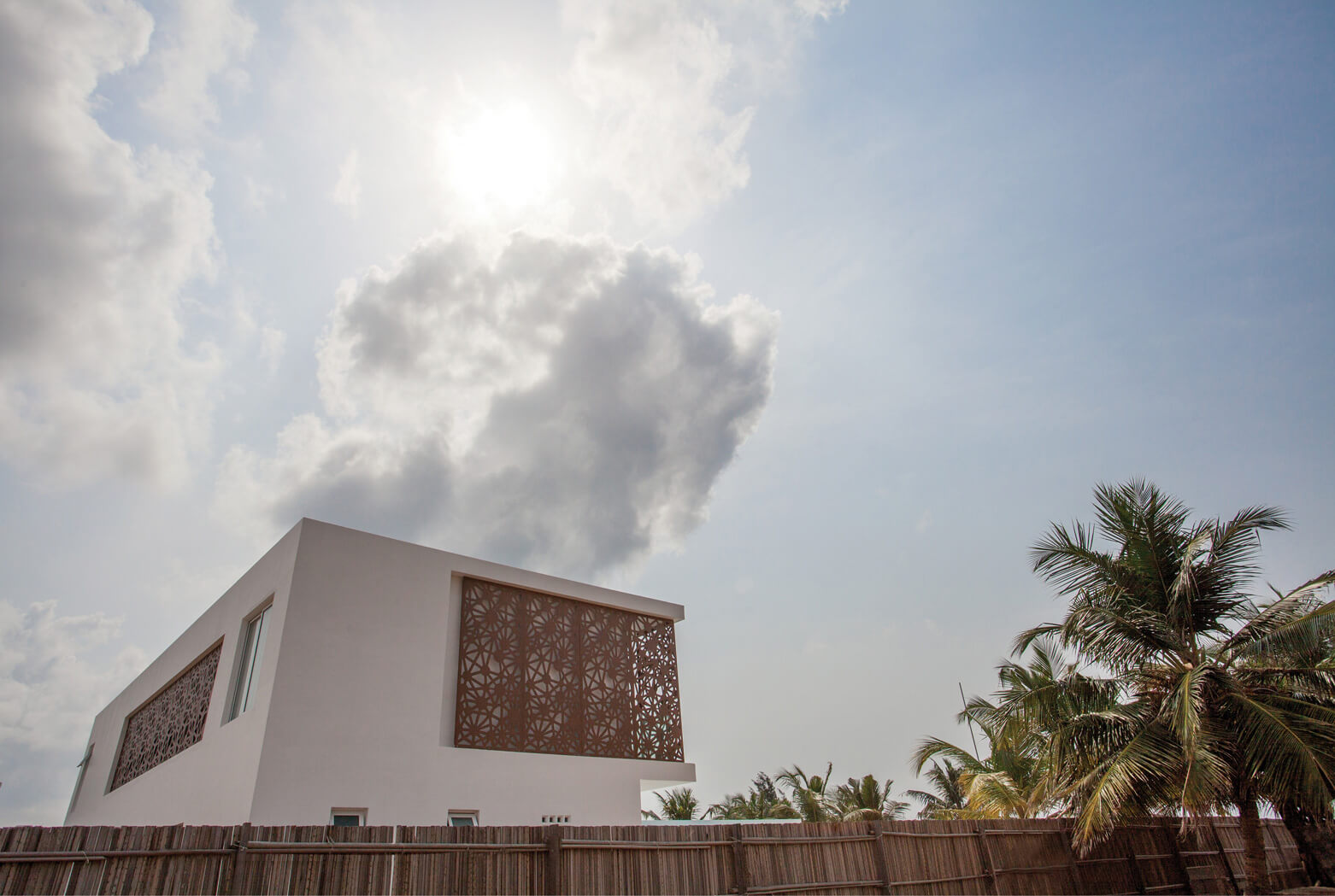 Dramatic sky above Sencillo Beach House, designed by Oshinowo Studio and led by Tosin Oshinowo. The building's distinct, angular architecture and patterned lattice screens stand out against a backdrop of fluffy white clouds and bright sunlight. Palm trees gently sway in the foreground, adding a natural and tropical touch to the modern, minimalist structure.