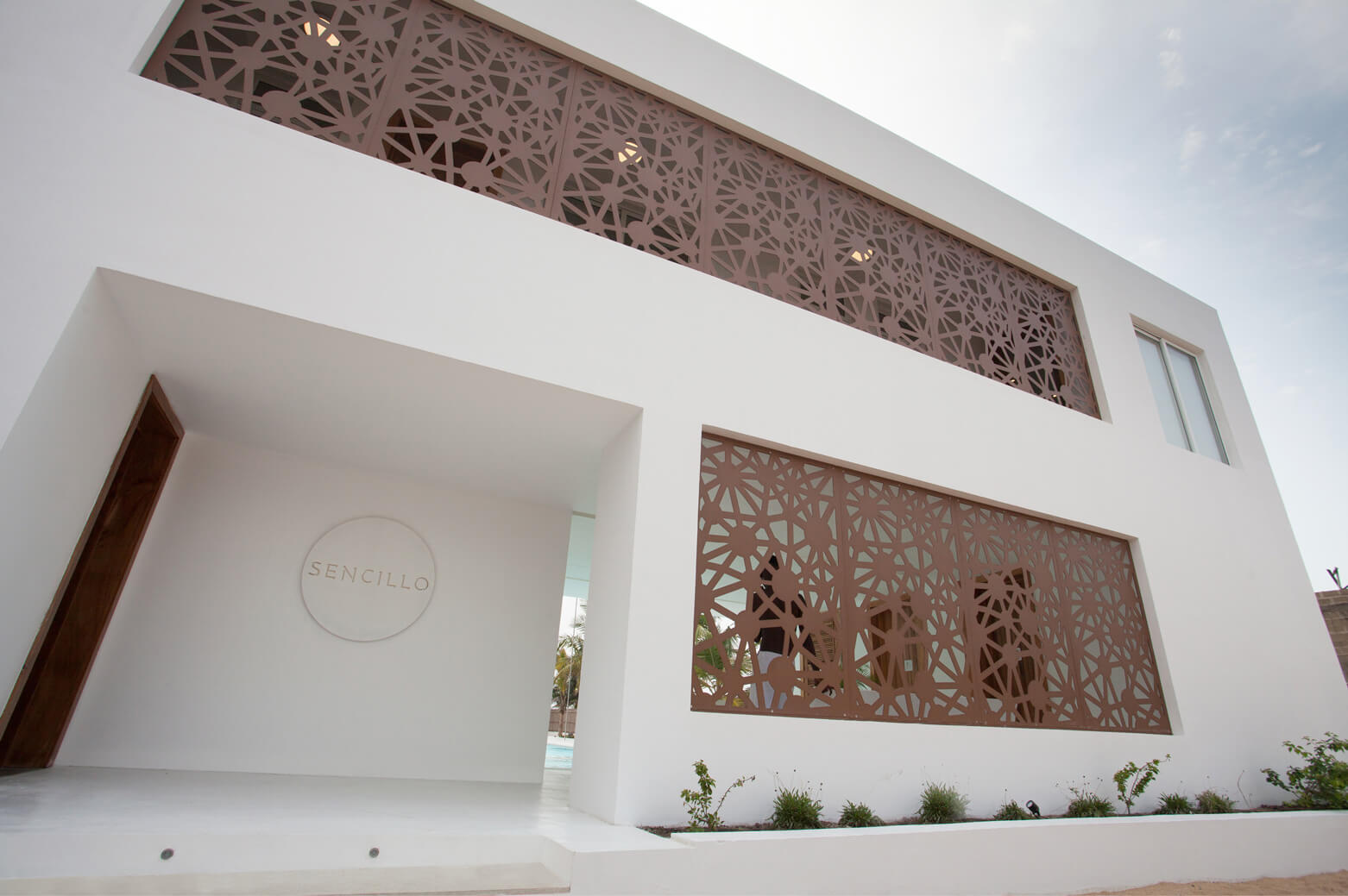 Close-up view of the entrance at Sencillo Beach House Lagos, designed by Oshinowo Studio and led by Tosin Oshinowo. The facade features a striking white geometric lattice design set against a crisp white background. The building is elegantly embossed in a circular design on the wall, which complements the minimalist architectural style. Nearby, small plants add a touch of greenery to the modern and serene environment