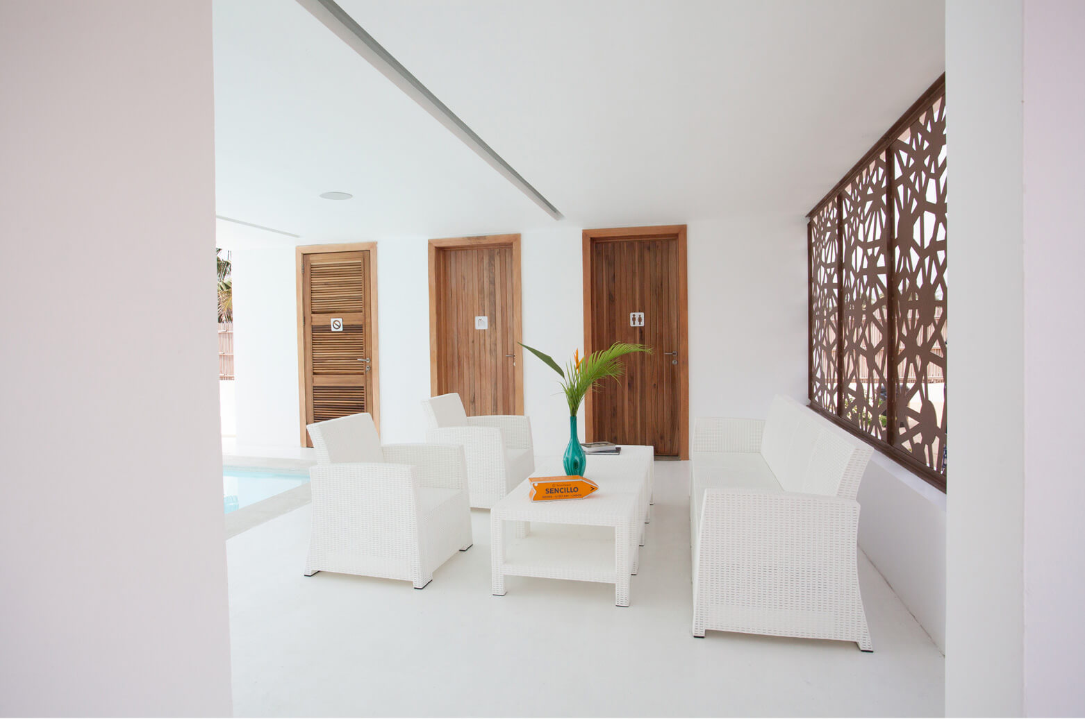 A stylish and serene seating area inside Sencillo Beach House, designed by Oshinowo Studio and led by Tosin Oshinowo. The space features minimalist white furniture arranged neatly on a sleek white floor, complemented by rich wooden doors and an intricate lattice window that allows natural light to filter through. A vibrant turquoise vase with lush green leaves adds a splash of color, enhancing the elegant and tranquil ambiance of the room.