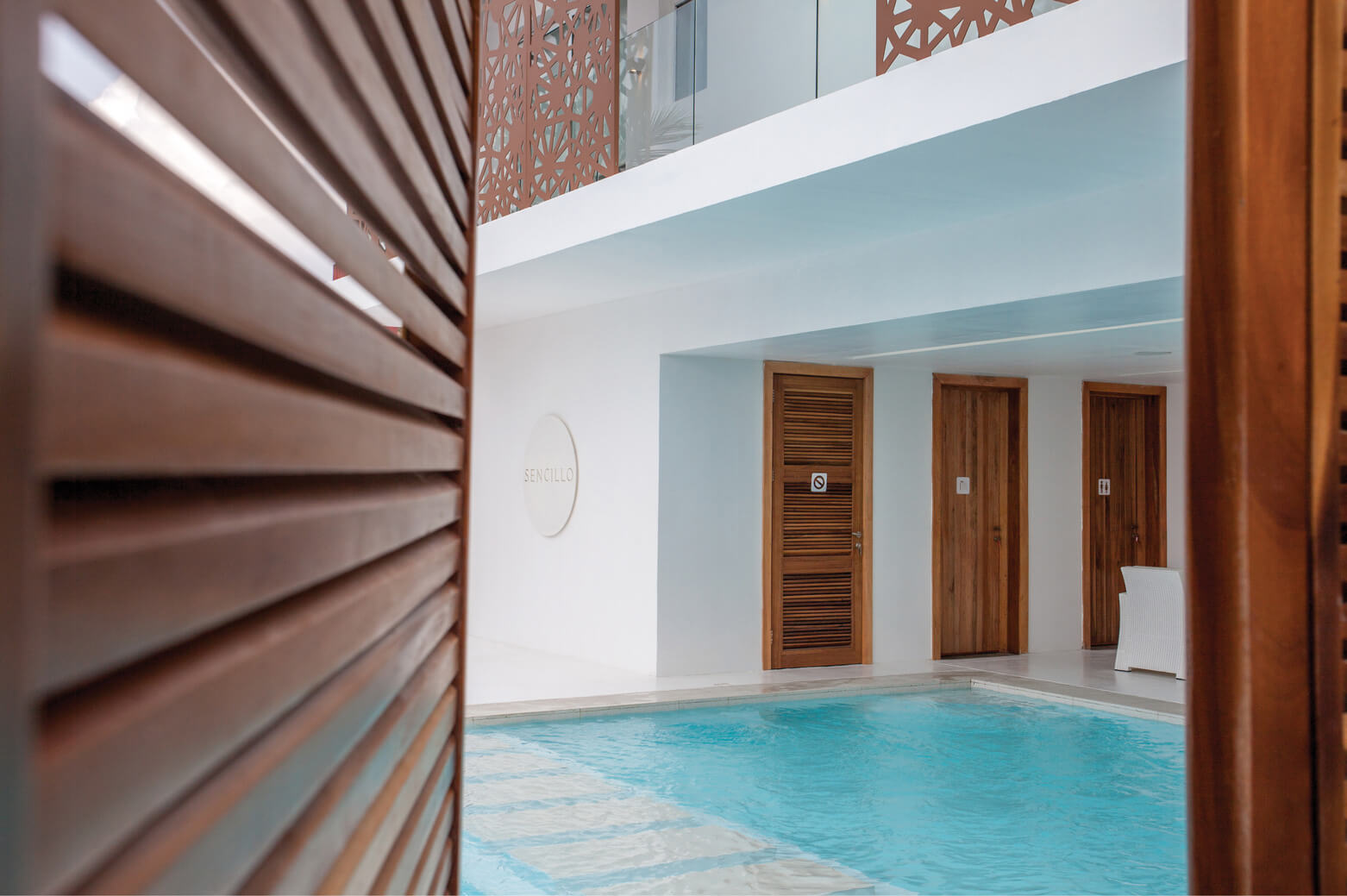 View through a wooden slatted partition to the tranquil pool area, designed by Oshinowo Studio and led by Tosin Oshinowo. The frame offers a peek into a serene setting featuring a clear blue pool, white architectural details, and wooden doors. The building’s name, ‘SENCILLO’, is elegantly displayed on a wall plaque, adding a sophisticated touch to the minimalistic yet inviting poolside ambiance.