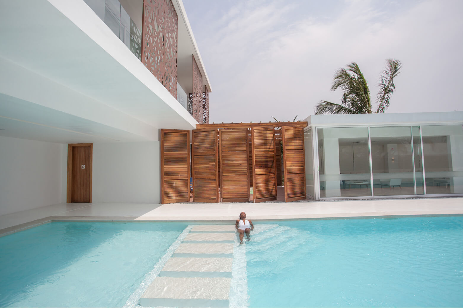 A leisurely day at the poolside, designed by Oshinowo Studio and led by Tosin Oshinowo. The pool’s crystal-clear turquoise waters invite relaxation, complementing the building’s white overhang and wooden slatted details. A child is seen enjoying the water, adding a playful element to the serene setting. The overall scene blends modern architecture with leisurely, resort-style living.