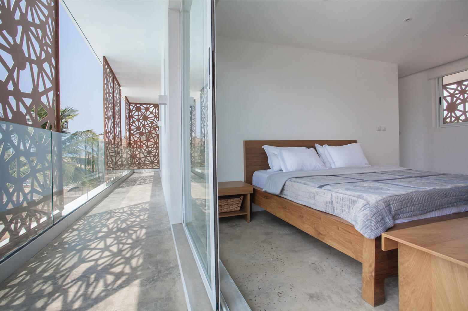 Bright and airy bedroom at Sencillo Beach House Lagos, designed by Oshinowo Studio and led by Tosin Oshinowo. This room features a large glass sliding door that opens to a balcony with intricate lattice railings, allowing for stunning views and light patterns created by the shadows of the lattice. A comfortable wooden bed with white bedding complements the room’s clean and minimalist aesthetic, harmoniously blending with the polished concrete flooring.