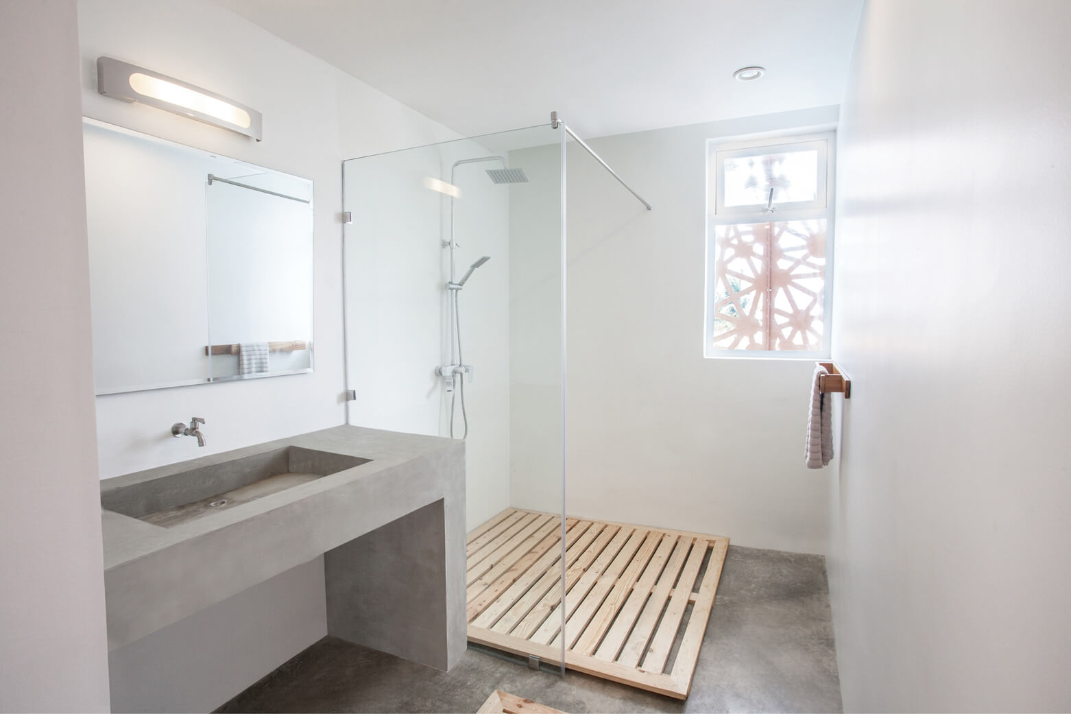 Modern minimalist bathroom at Sencillo Beach House Lagos, designed by Oshinowo Studio and led by Tosin Oshinowo. The space is sleek and functional, featuring a long concrete sink counter and a spacious walk-in shower enclosed by clear glass. A unique wooden slatted floor mat in the shower area adds a natural touch. The room is brightly lit by natural light filtering through a window that showcases the distinctive patterned lattice design, visible outside.