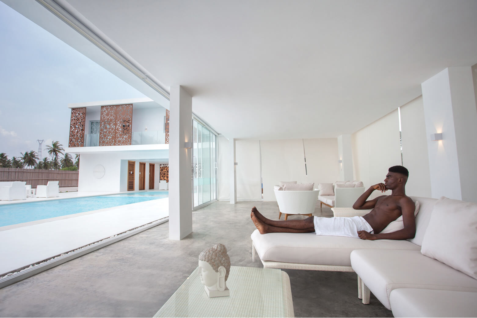 A serene indoor-outdoor living space at Sencillo Beach House Lagos, designed by Oshinowo Studio and led by Tosin Oshinowo. The area features an open layout with large sliding glass doors offering a view of the pool and the distinctive lattice-paneled building facade. A young man relaxes on a minimalist white couch, embodying the tranquil, luxurious atmosphere of the setting.