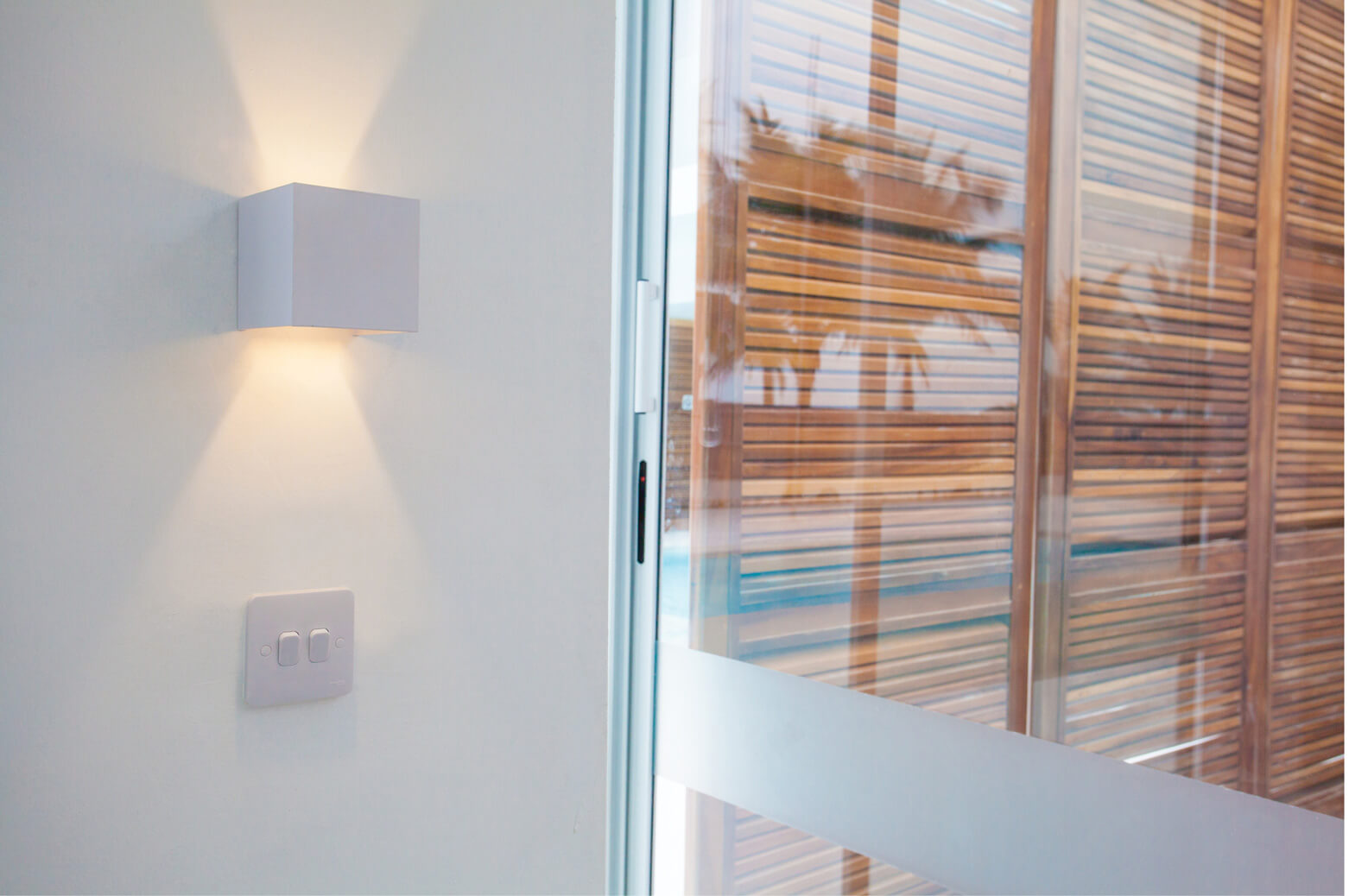 A detail-oriented interior shot at Sencillo Beach House, designed by Oshinowo Studio and led by Tosin Oshinowo. The image captures a sleek, modern wall light casting a bright, angular glow against a smooth white wall. Adjacent, a glass door provides a glimpse of wooden blinds and tropical greenery outside, creating a play of light and shadows that adds depth and warmth to the minimalist design.