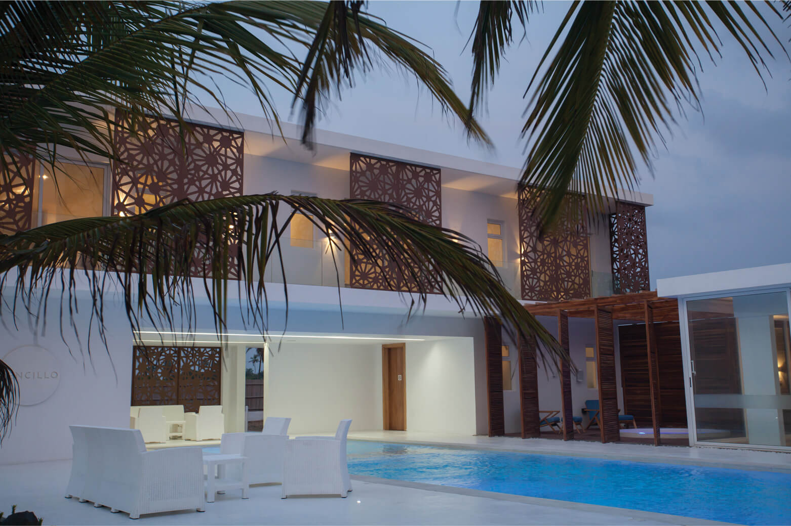 An evening view of Sencillo Beach House Lagos, designed by Oshinowo Studio and led by Tosin Oshinowo. The scene captures the warm glow of interior lights shining through the unique, geometric lattice panels that adorn the building's exterior. Palm fronds frame the foreground, enhancing the tropical ambiance. A serene pool reflects the subtle lighting, surrounded by white outdoor furniture, providing a calm and luxurious setting.