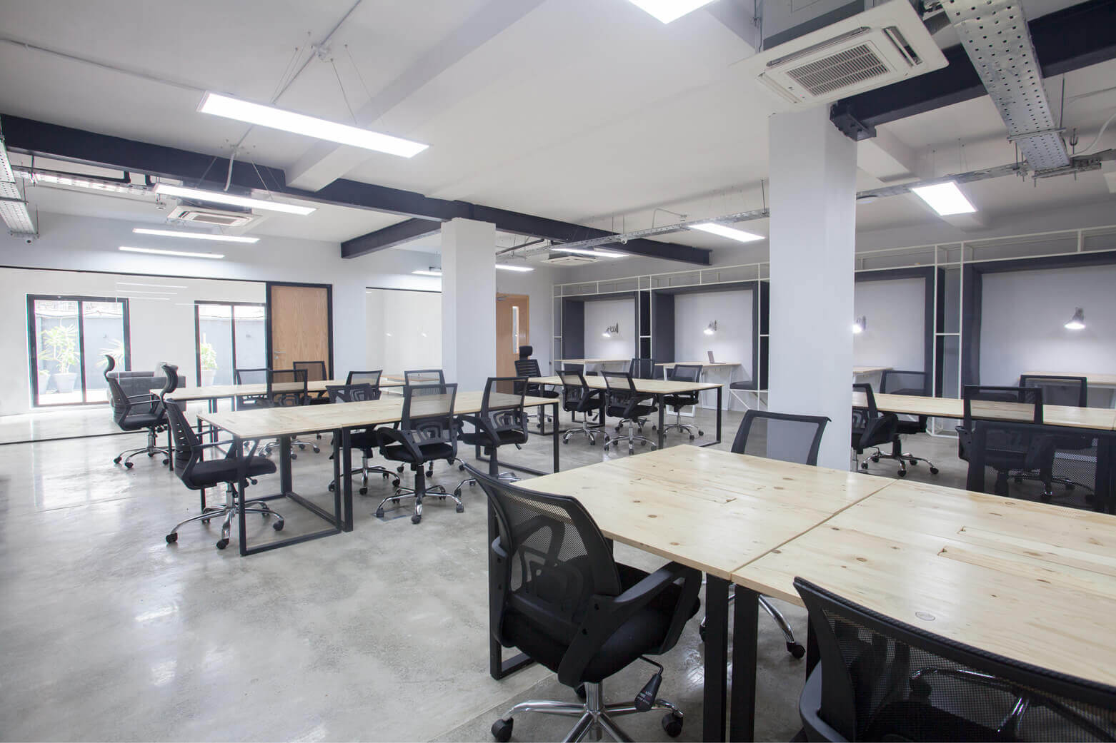 A spacious, well-lit modern office environment, possibly designed by Oshinowo Studio, a leading architectural design studio in Lagos, led by Tosin Oshinowo, and energized by Rensource. The interior features multiple light wooden tables paired with ergonomic black chairs, set on a polished concrete floor that reflects the ambient lighting. Overhead, strip lighting and air conditioning units are installed into the exposed ceiling, contributing to a contemporary industrial aesthetic. The space, defined by white columns and framed by large windows, exudes a clean and professional atmosphere.