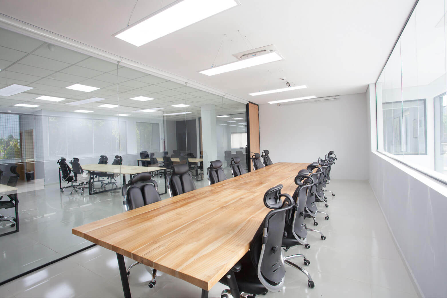 This is an image of a bright and modern office interior, likely designed by the leading architectural design studio in Lagos, Oshinowo Studio, led by Tosin Oshinowo, and powered by Rensource energy solutions. The office space is furnished with light wood conference tables lined with sleek black office chairs. The room is well-lit by natural light from the windows along the wall and complemented by overhead fluorescent lights, creating a productive ambiance. The floor is polished to a shine, reflecting the clean and minimalistic aesthetic of the room.