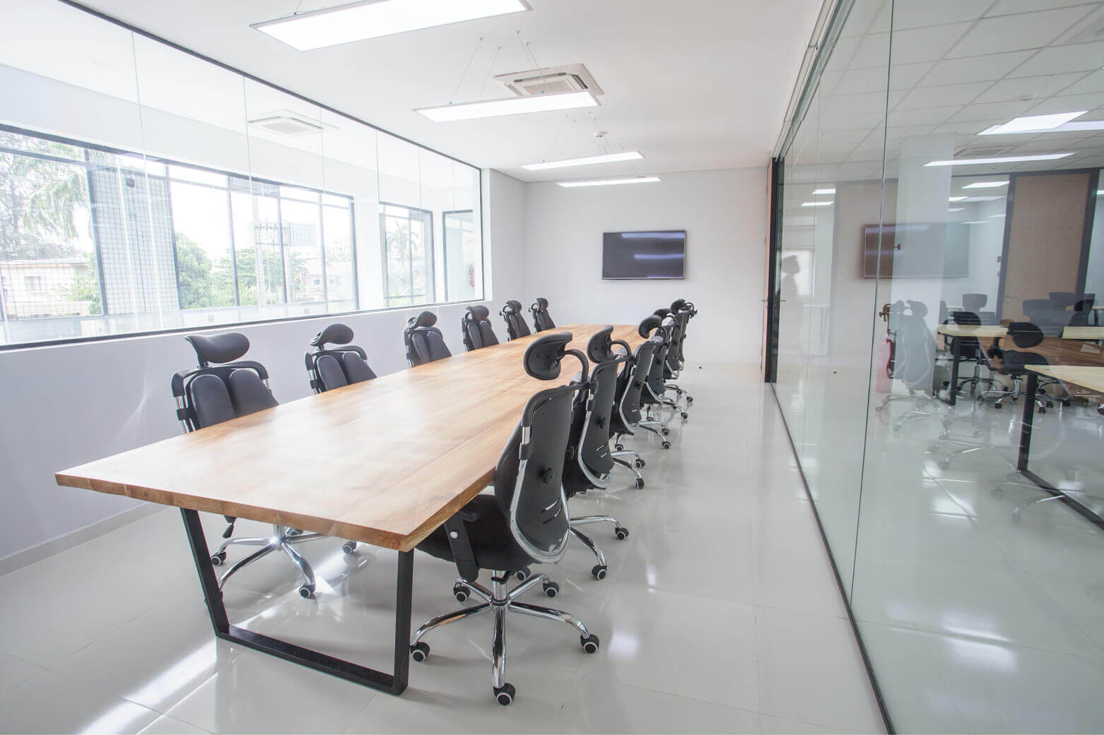Stylish and functional meeting room at the leading architectural design studio in Lagos, Oshinowo Studio, led by Tosin Oshinowo, featuring a long wooden table with sleek black legs, complemented by high-backed rolling office chairs. The room is framed by floor-to-ceiling glass walls that allow for a flood of natural light, and a flat-screen TV mounted on the wall for presentations, reflecting the studio's modern and collaborative work ethos.