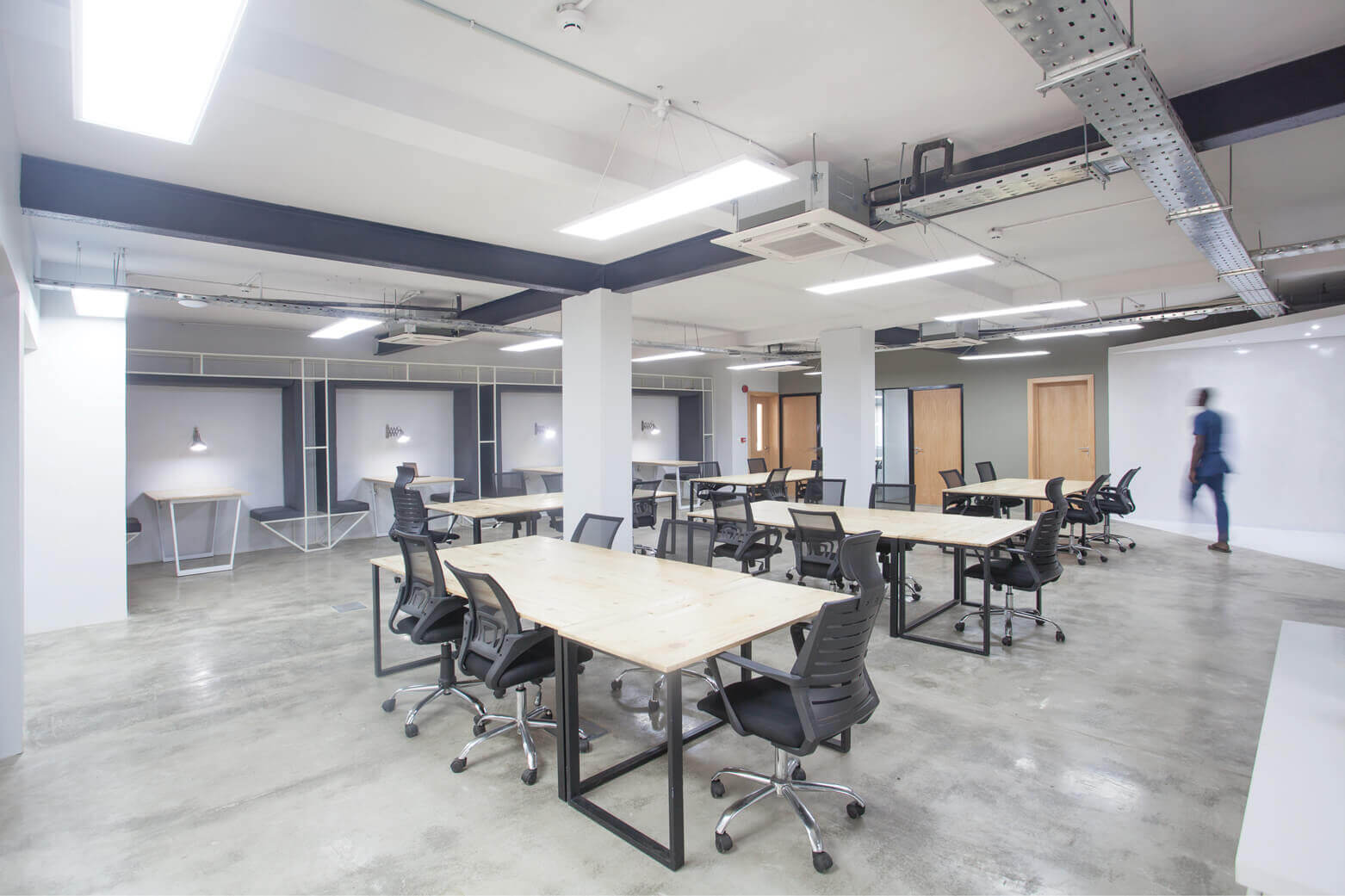 This image presents a modern office space that embodies the design expertise of Oshinowo Studio, a leading architectural design studio in Lagos. The room is airy and bright, with natural light streaming through large windows and complementing the industrial-style lighting from overhead fixtures. Several wooden tables with black office chairs are arranged throughout the space, set against a backdrop of white walls and grey concrete flooring. In the background, a figure in blue is blurred in motion, adding a dynamic element to the otherwise still and orderly office setting.