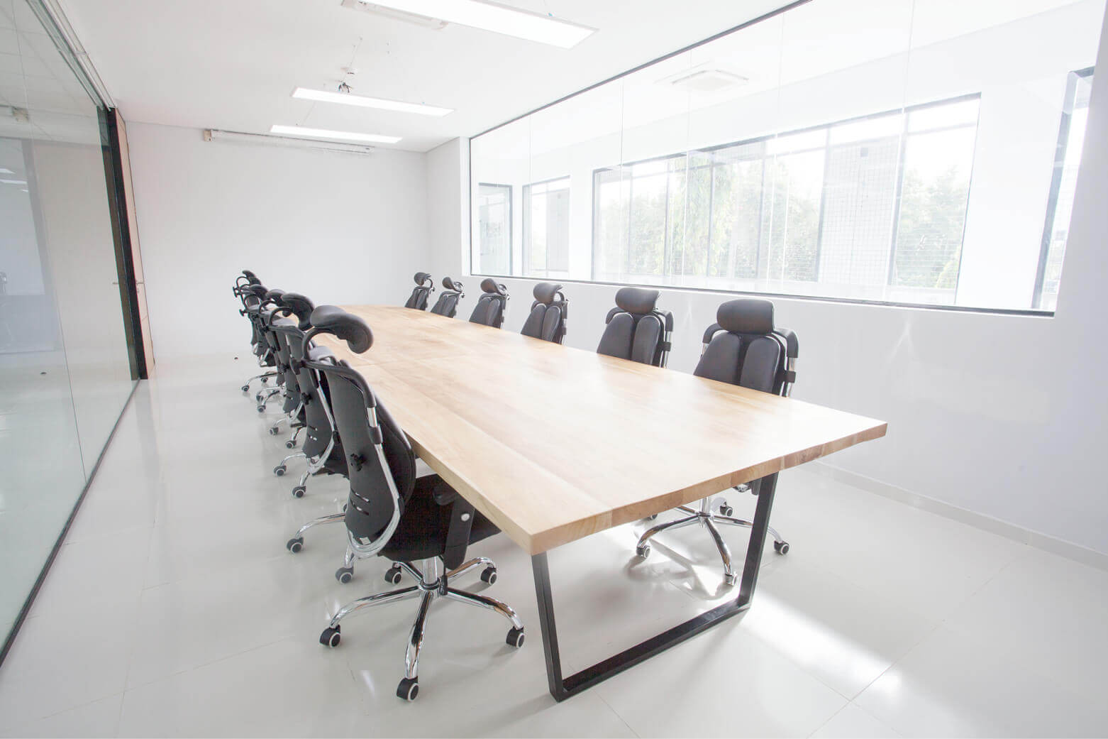 A minimalist and contemporary conference room, possibly designed by Oshinowo Studio, a leading architectural design studio in Lagos, led by Tosin Oshinowo. The room is dominated by a large wooden table with a polished natural finish, surrounded by ergonomic black office chairs on casters. The space is bright and airy, illuminated by natural light streaming in from the large windows on the side and complemented by recessed ceiling lights. The room's clean lines and uncluttered design create an atmosphere conducive to focus and collaboration.
