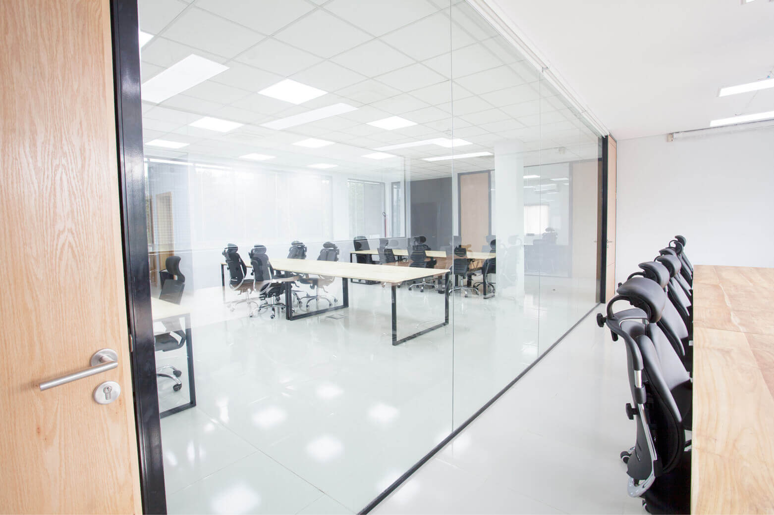 Modern conference room at the leading architectural design studio in Lagos, Oshinowo Studio, led by Tosin Oshinowo, featuring a clean and open design with light wooden tables and black office chairs. The room is characterized by its reflective white floor, drop ceiling with recessed lighting, and wooden doors that provide a warm contrast to the otherwise monochrome color scheme. A person in motion adds a dynamic element to the scene, indicating the collaborative and active environment of the studio.