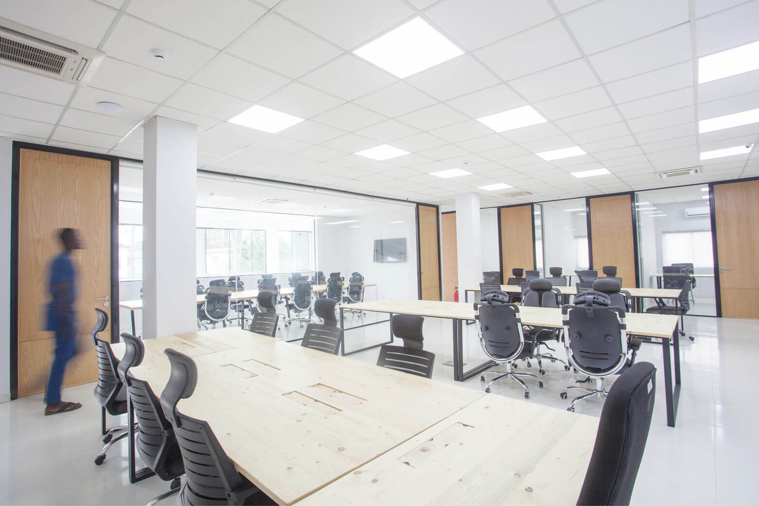 Modern and airy meeting area in the leading architectural design studio in Lagos, Oshinowo Studio, led by Tosin Oshinowo. It features a bright, open space with large tables with natural wooden surfaces and stylish black chairs. The blurred motion of a person walking through adds dynamism to the scene, set against a backdrop of clean white walls, wooden doors, and a grid of ceiling lights that provide a well-lit and inviting environment for collaboration and design.