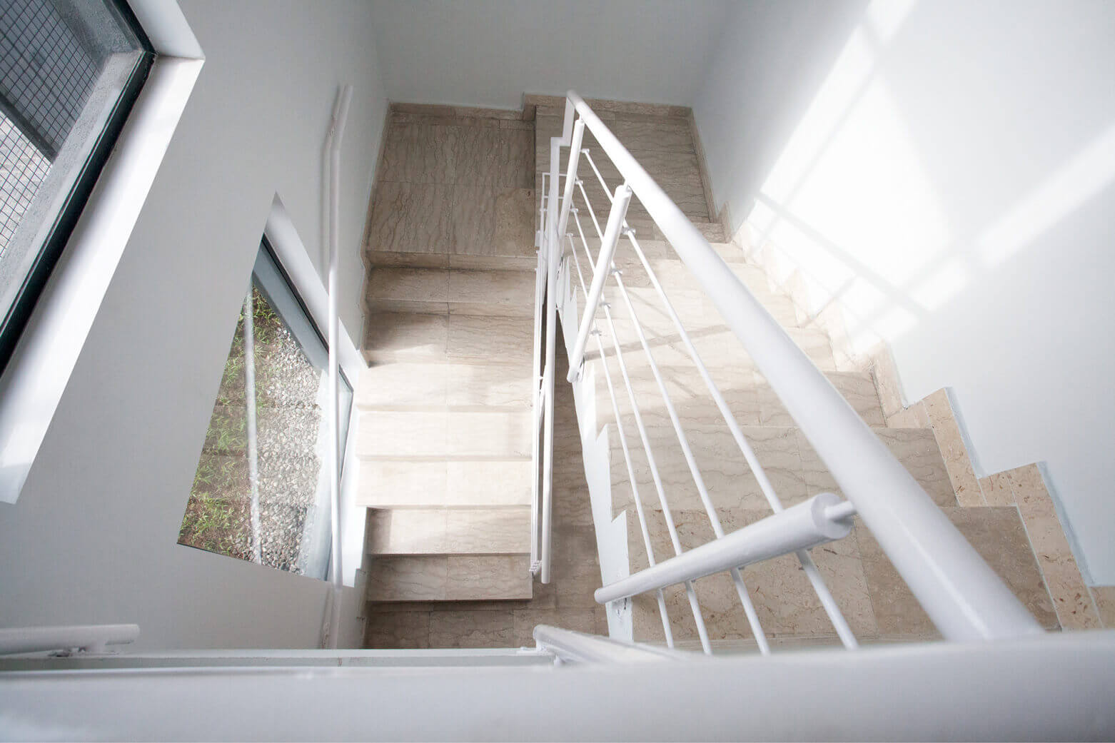 A perspective view of a modern staircase inside a building designed by Oshinowo Studio, a leading architectural design studio in Lagos. The staircase, composed of light marble steps, is accentuated by white-painted steel railings and bright natural light streaming through the geometrically shaped windows. The purity of the white walls and the transparency of the space embody the studio's minimalist and contemporary design ethos.