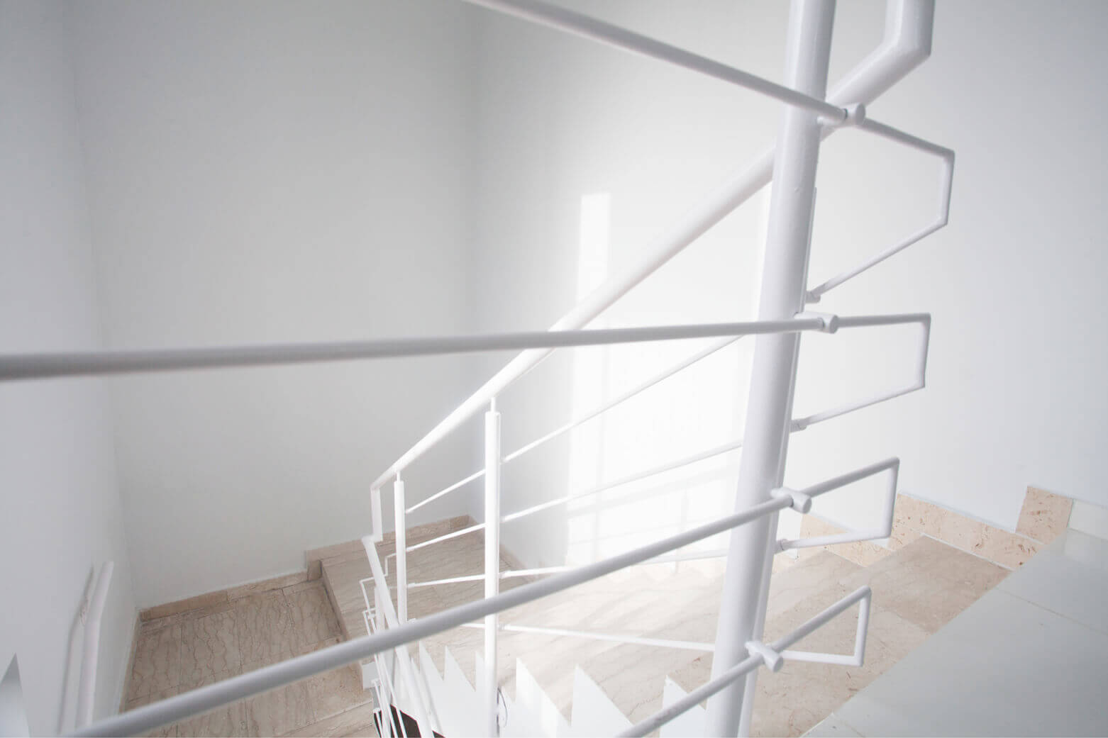An image capturing the serene and minimalist design of a staircase within a building created by Oshinowo Studio, a leading architectural design studio in Lagos. The photograph shows a view from the top of the staircase, looking down on the clean lines of the white balustrades and the light that floods in from a large window. The steps are made of a light-colored marble, and the space is characterized by its bright, airy feel and modern aesthetic.