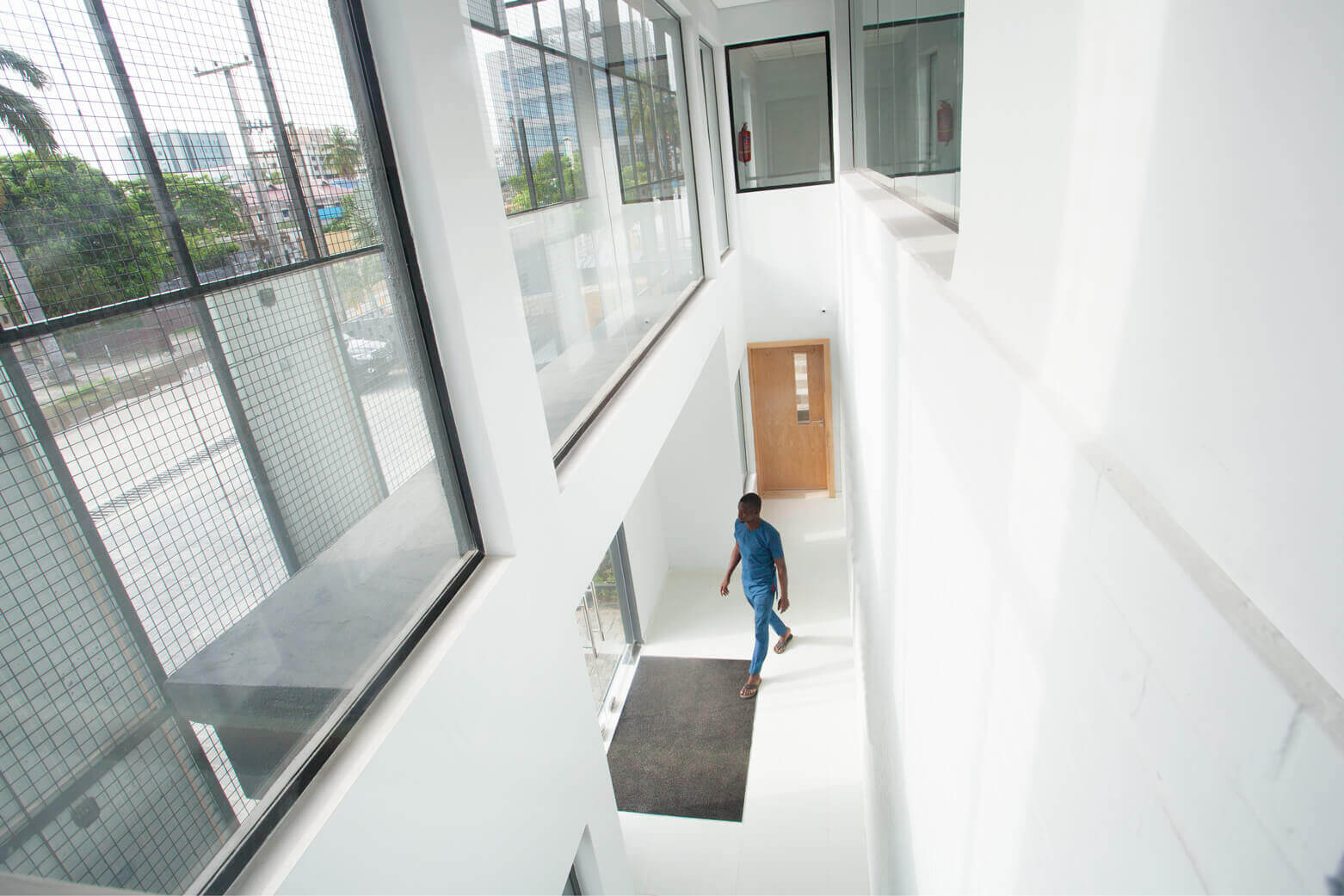 A man in blue attire walks through a sunlit corridor of a modern building, possibly a design by Oshinowo Studio, a leading architectural design studio in Lagos. The corridor features clean lines, a white palette, and large windows that provide a view of the city and allow natural light to flood the space. The design emphasizes transparency and brightness, contributing to an open and airy atmosphere within the building.