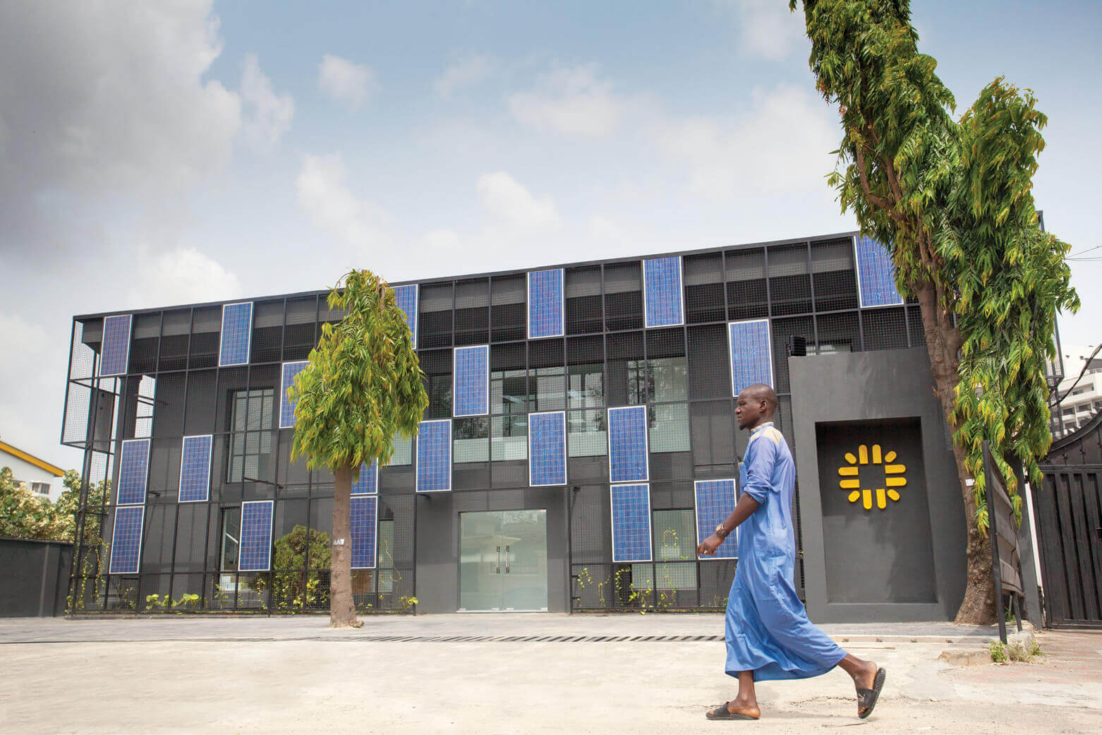 A man in traditional blue attire walks past a modern building clad with blue solar panels on its façade in Lagos. The building, featuring a two-story design with a geometric structure, is branded with the Rensource logo, indicating a sustainable energy solution. In the foreground, lush greenery accentuates the building's commitment to eco-friendliness, showcasing the innovative work of the leading architectural design studio, Oshinowo Studio, led by Tosin Oshinowo