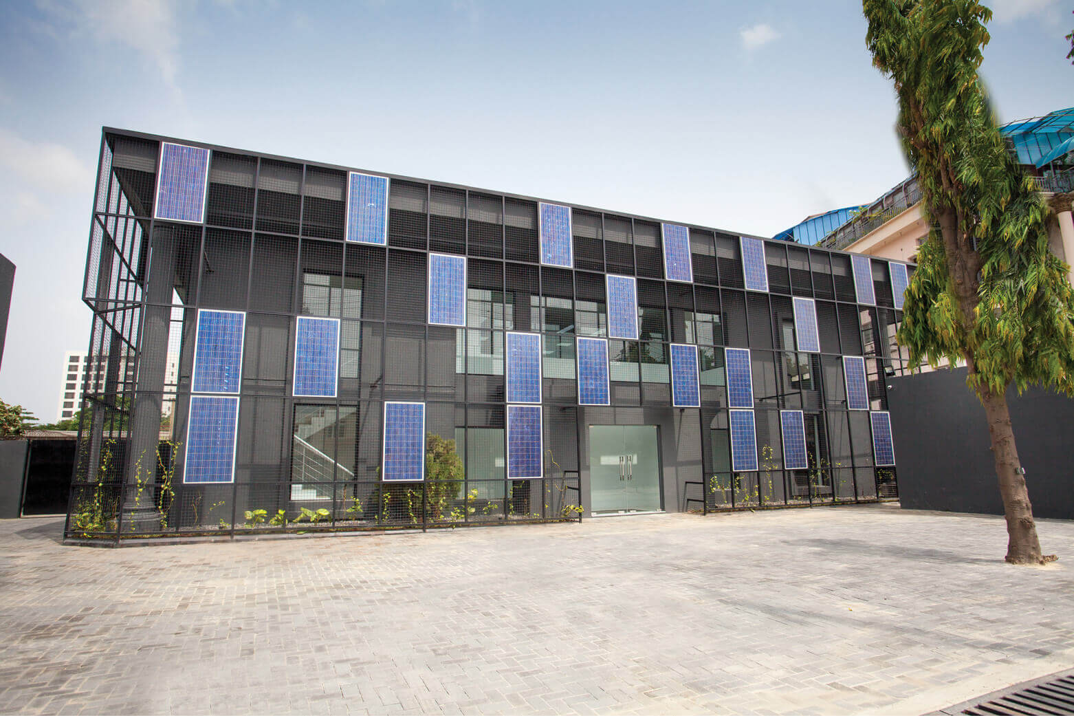 A modern, eco-friendly building facade featuring a grid of blue solar panels against a black mesh exterior, located in Lagos. The clear sky above contrasts with the building's energy-efficient design, a project by Rensource. This innovative structure, exemplifying the sustainable architectural prowess of the leading design studio, Oshinowo Studio, led by Tosin Oshinowo, is surrounded by a paved area and adorned with green plants at the base, enhancing its blend with the environment.