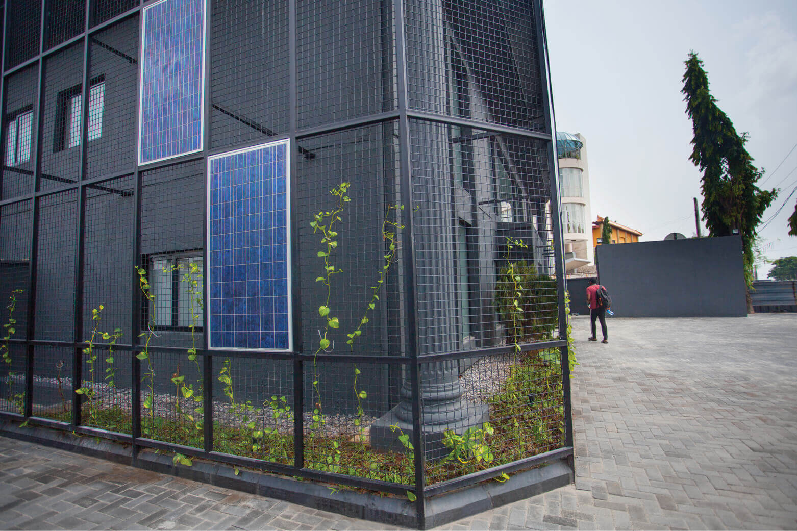 A person in a red shirt and dark pants is seen from behind, walking along the paved area adjacent to an innovative black building adorned with blue solar panels and a protective mesh. Green vines begin to climb the metal grid, hinting at the integration of nature into the design. This energy-conscious edifice in Lagos is a distinctive project by Rensource, reflecting the forward-thinking environmental solutions championed by the leading architectural design studio, Oshinowo Studio, led by Tosin Oshinowo.