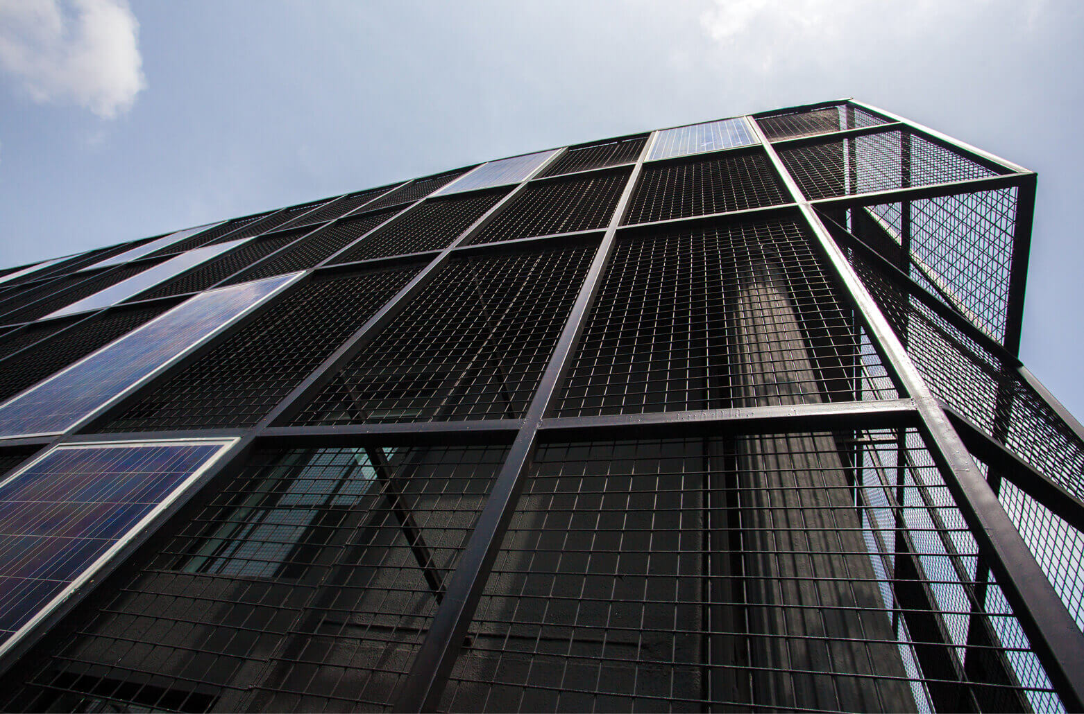 A dramatic upward view of a cutting-edge, multi-story building clad in a mesh-like material with embedded blue solar panels. This architectural marvel, likely a collaborative effort by Rensource for sustainable energy solutions and Oshinowo Studio, a leading architectural design studio in Lagos, led by Tosin Oshinowo, stands against a bright sky with scattered clouds. The design emphasizes verticality and environmental responsibility, signaling a forward-thinking approach to urban architecture.