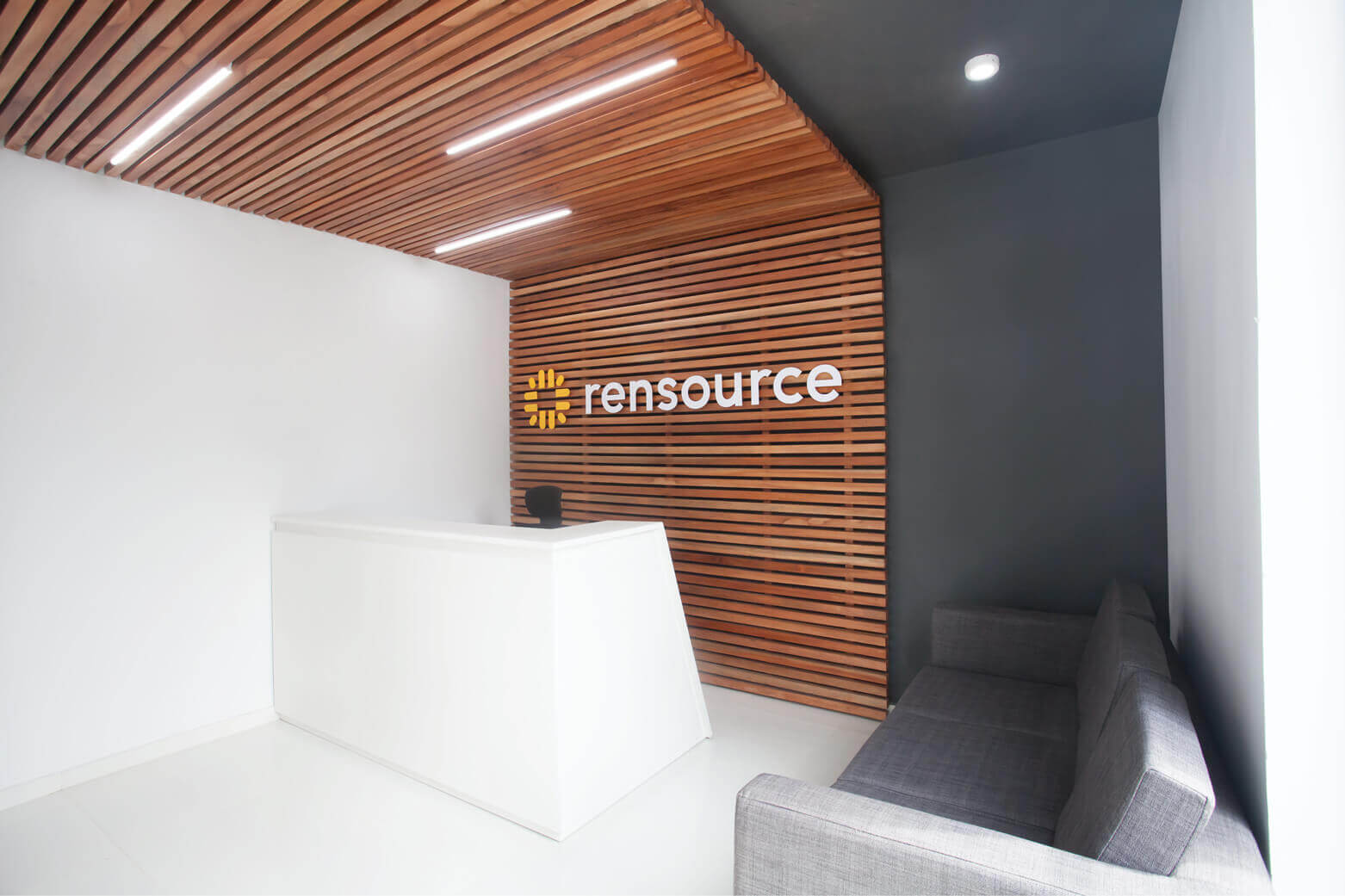 The interior of a sleek and modern office reception area, possibly a collaboration between Rensource and Oshinowo Studio, a leading architectural design studio in Lagos, led by Tosin Oshinowo. The reception desk is a minimalist white counter positioned in front of an accent wall with horizontal wooden slats where the Rensource logo is prominently displayed. Above, linear lights are set into a wooden ceiling, casting a warm glow. To the right, a plush gray couch offers a comfortable waiting area, complementing the monochrome palette of the walls and floor.