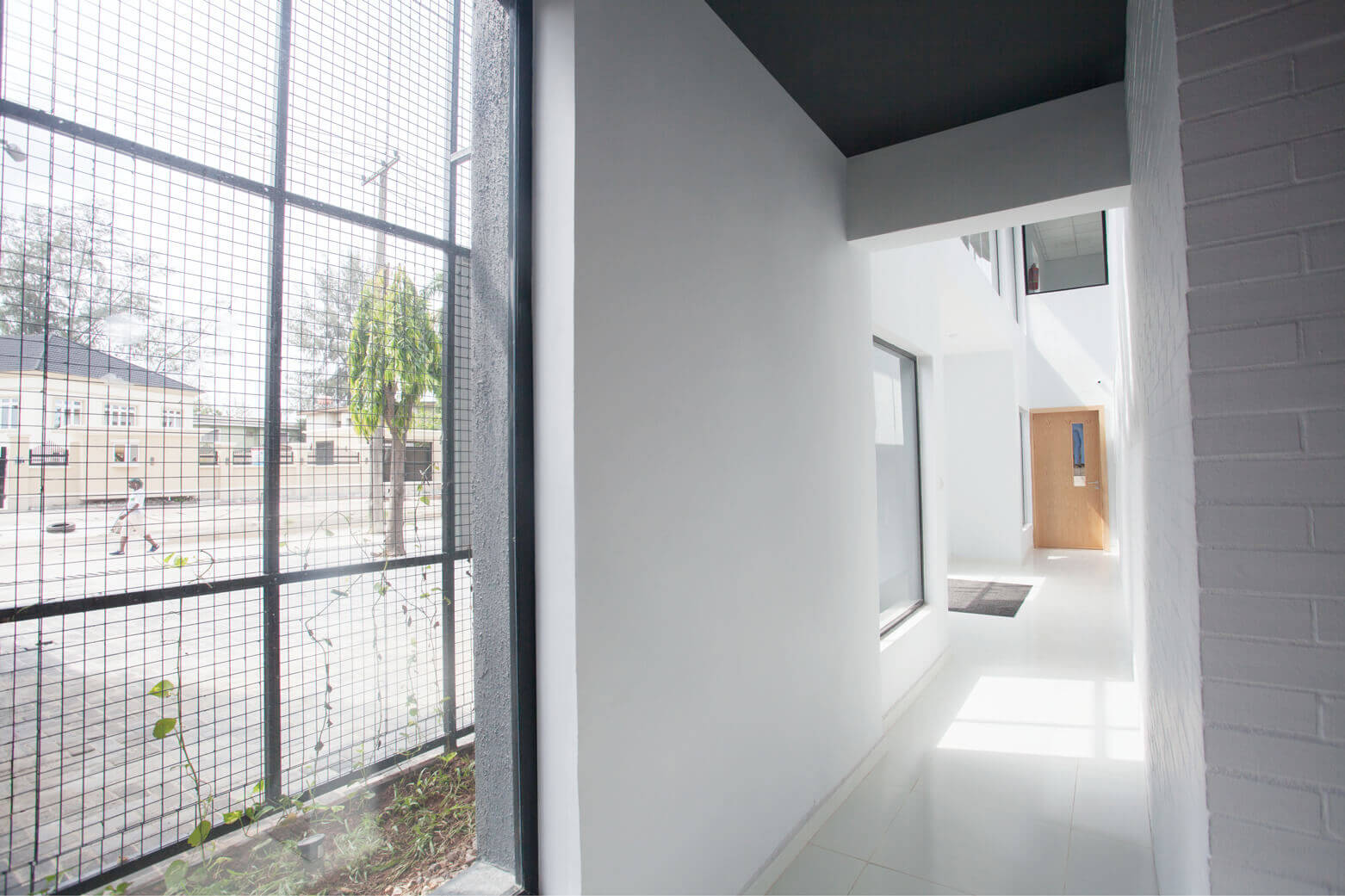 View through the corridor of a leading architectural design studio in Lagos, Oshinowo Studio, highlighting the sleek, modern design with white walls and polished floors. The corridor is framed by large windows covered with protective mesh, offering a glimpse of the greenery and urban scenery outside, while ensuring a bright, naturally lit pathway leading to wooden doors that signify the entrance to creative workspaces.