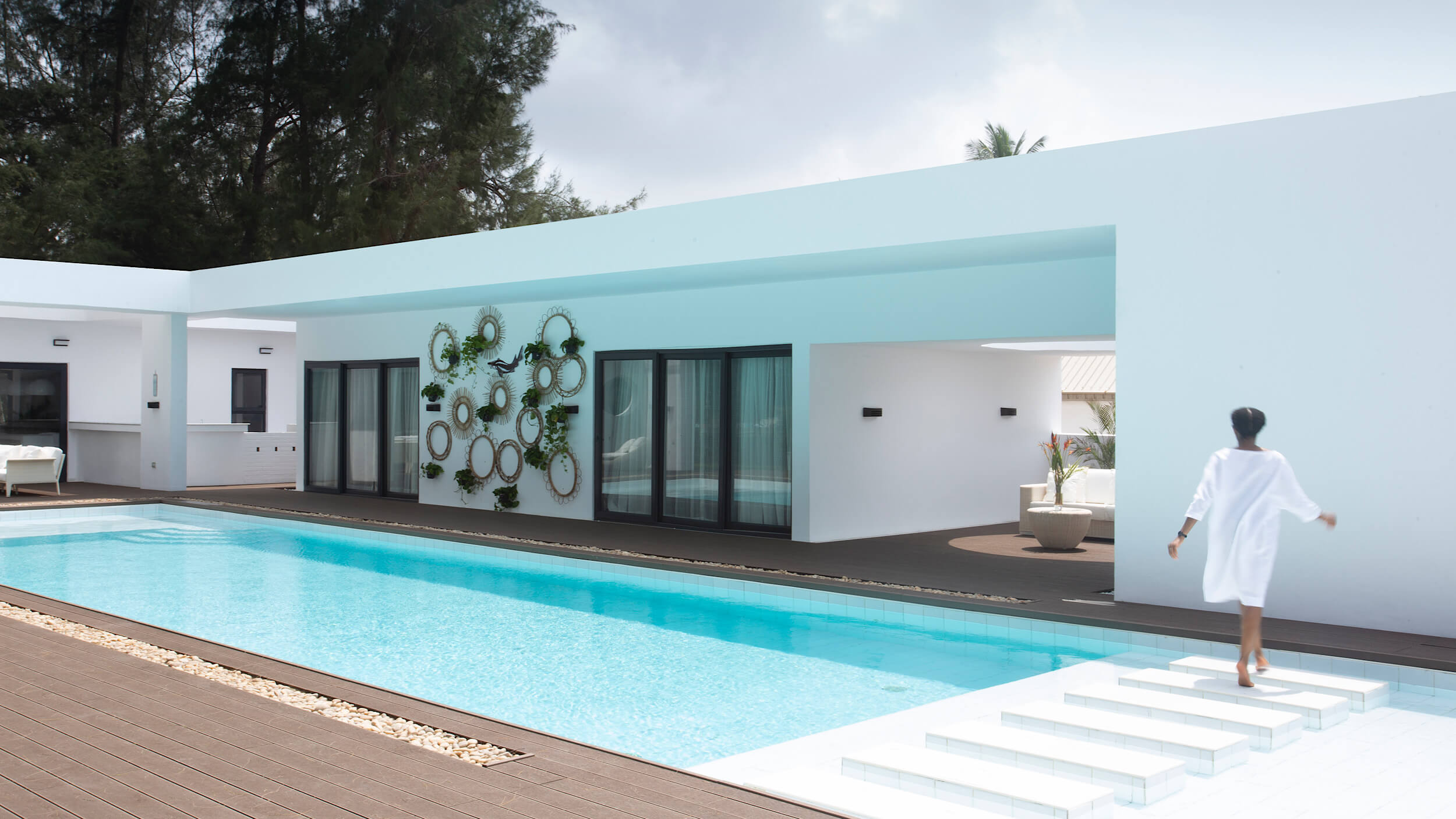 A luxurious white villa with a rectangular swimming pool in the foreground, designed by Oshinowo Studio and led by Tosin Oshinowo. The villa features clean, minimalist architecture with a large white wall and a soft blue overhang. On one side, a series of mirrors arranged as wall art enhances the building's aesthetic. A woman in a white robe is captured in motion, walking across white steps that lead down to the pool, surrounded by lush green trees in the background.
