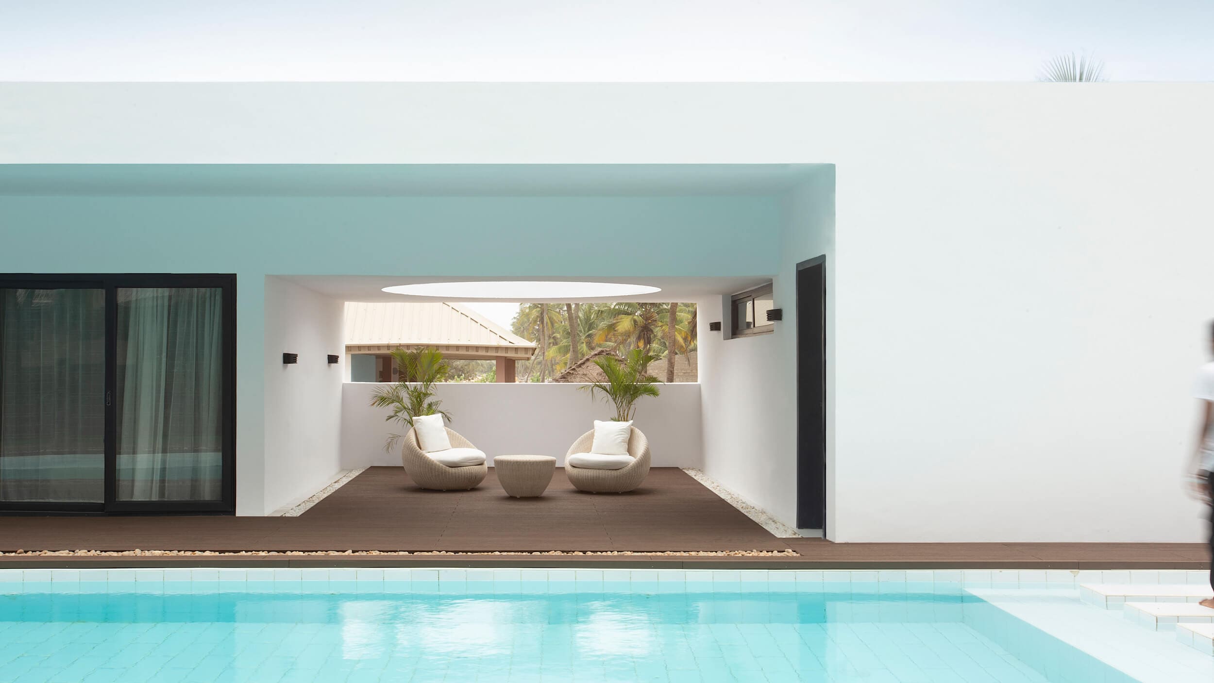 A serene view of an outdoor relaxation area at a luxurious villa, designed by Oshinowo Studio and led by Tosin Oshinowo. The space is framed by a large turquoise beam extending across the top. Below, two stylish white woven chairs with matching footrests sit on a dark brown deck, flanked by potted plants, creating an inviting lounge area. The deck overlooks a bright turquoise swimming pool in the foreground. Part of a person in a white robe is visible on the right edge, suggesting movement. The backdrop features tall palm trees and a clear sky.