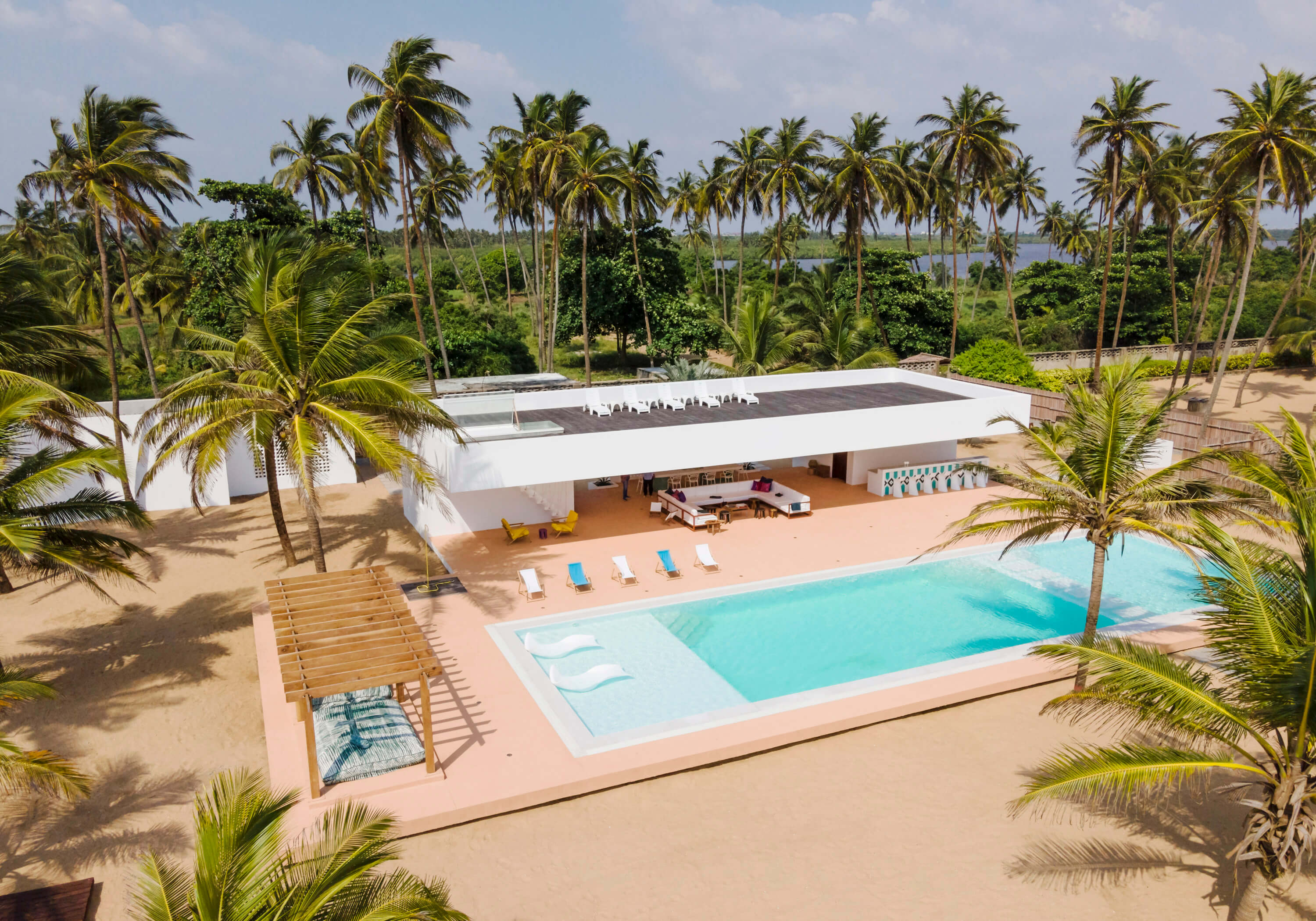 Tosin Oshinowo led the design and execution of the aerial view of the Coral Pavilion, a modern, flat-roofed villa with solar panels, designed by the leading architectural design studio in Lagos, Oshinowo Studio. The villa is nestled among lush palm trees, with a large rectangular swimming pool surrounded by pinkish sand-colored poolside area with loungers, a covered patio with seating, and an open-air dining setup. The ocean is visible in the background, suggesting a coastal location.