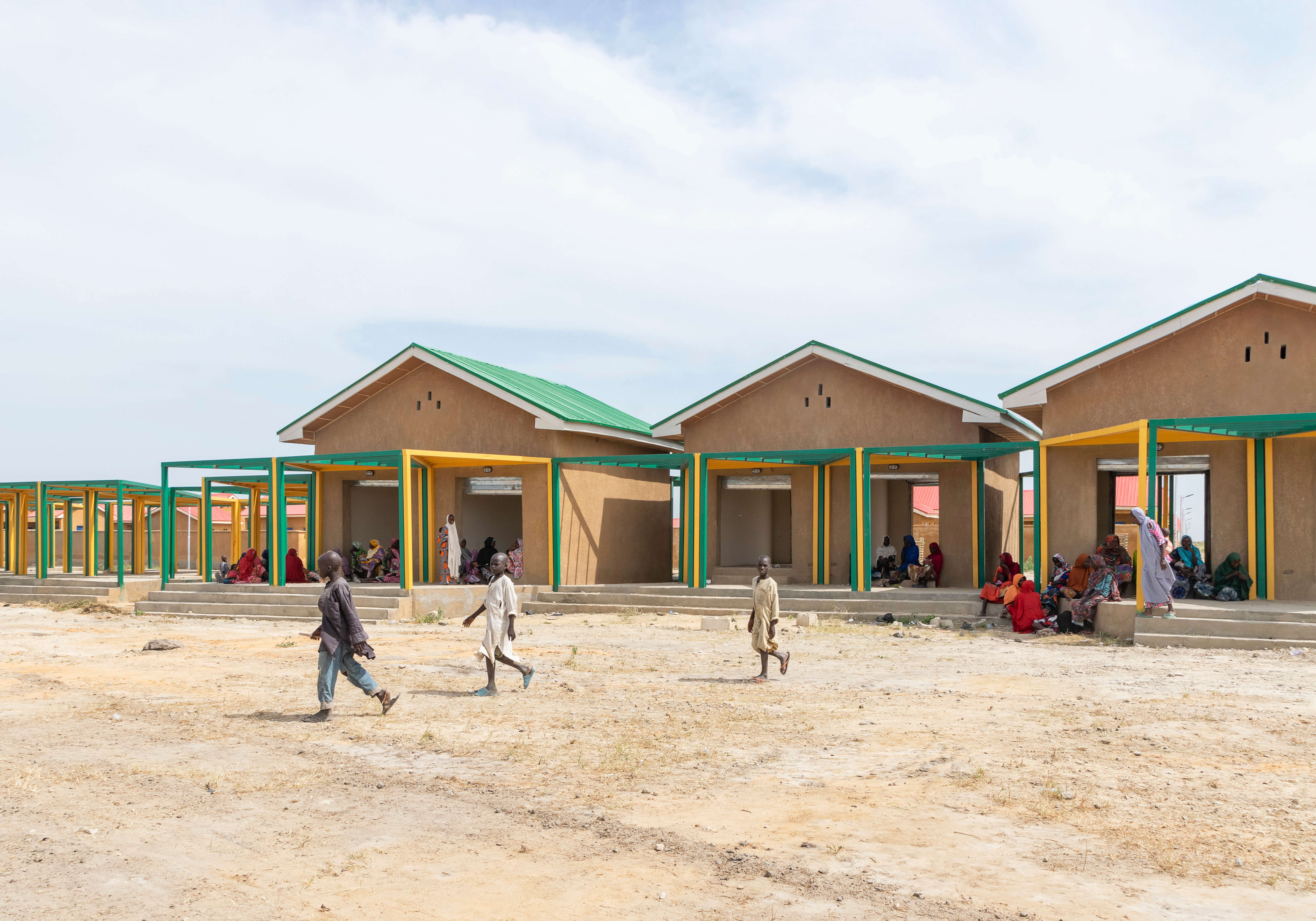 A view of a community marketplace in a sandy, open area, featuring design elements from Oshinowo Studio, a leading architectural design studio in Lagos, led by Tosin Oshinowo. The market includes several ochre-colored buildings with green metal roofs, complemented by yellow and green pillars. Local people are gathered in small groups, seated on benches and ledges under the shade of the overhanging roofs, while children play in the foreground, animating the serene communal space.