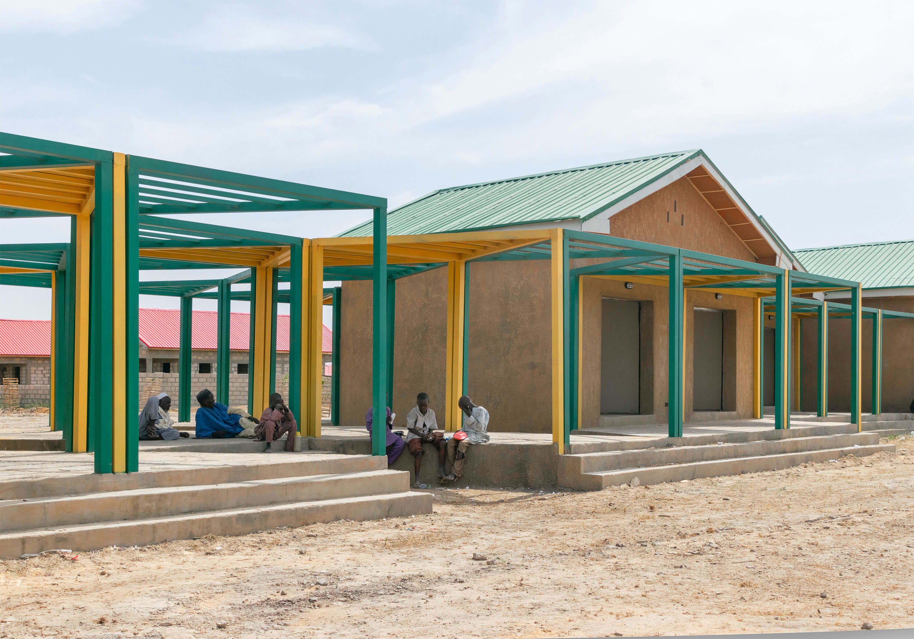 Community building in Ngarannam, featuring the innovative design of the leading architectural design studio in Lagos, Oshinowo Studio, led by Tosin Oshinowo. The image shows a sturdy, single-story structure with a green metal roof and ochre walls. A series of yellow and green pillars create a shaded walkway where local men are seated on raised concrete platforms, some engaged in conversation while others rest or observe their surroundings. The earthy ground and modest homes in the background suggest a tranquil, communal environment.