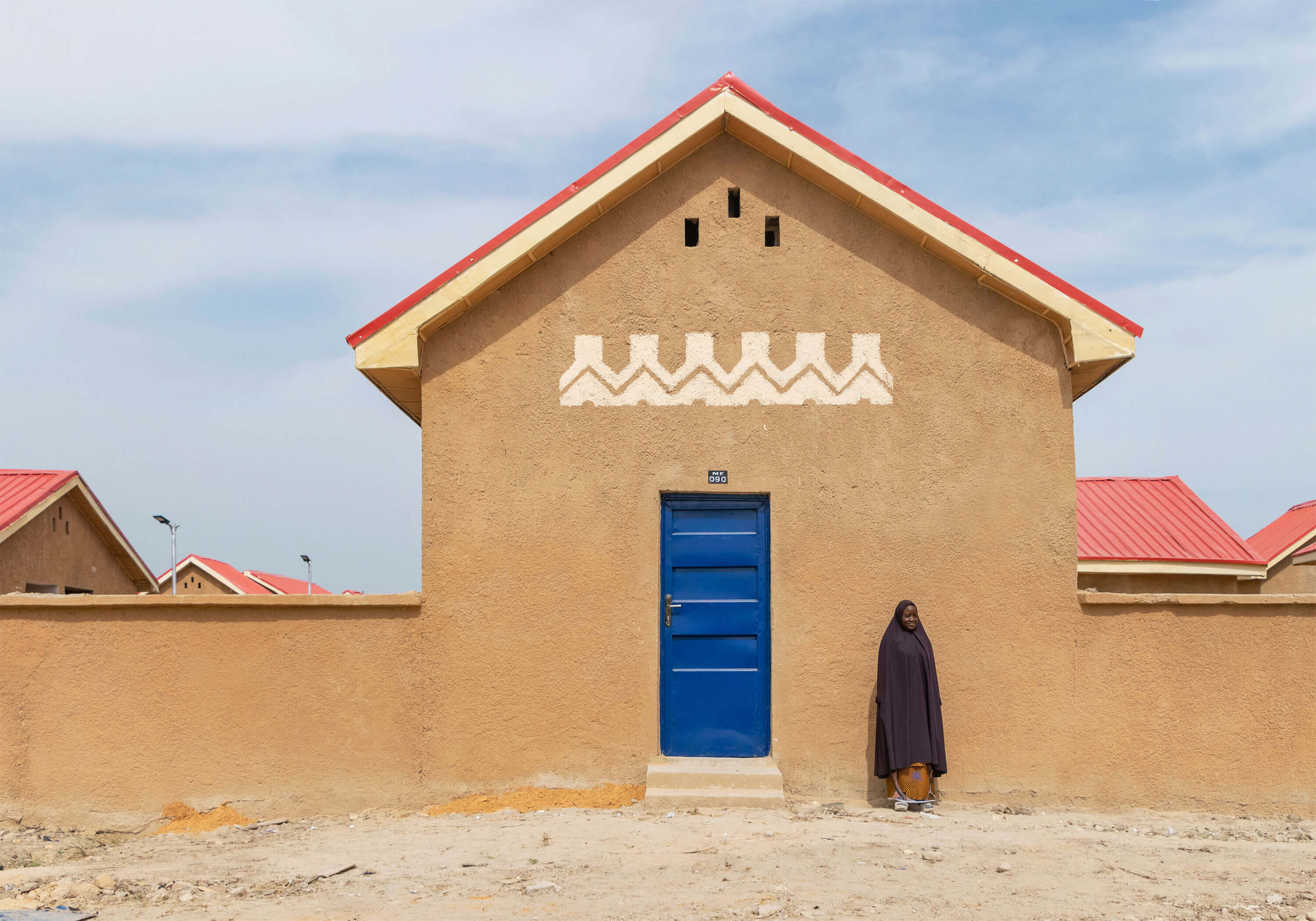 In Ngarannam, a woman stands in front of a house featuring the distinct aesthetic of Oshinowo Studio, a leading architectural design studio in Lagos, led by Tosin Oshinowo. The house's ochre walls are adorned with traditional white patterns, complementing the vivid blue door and red metal roof. This simple yet striking facade captures the essence of local design elements and color palettes, creating a sense of cultural identity and place.
