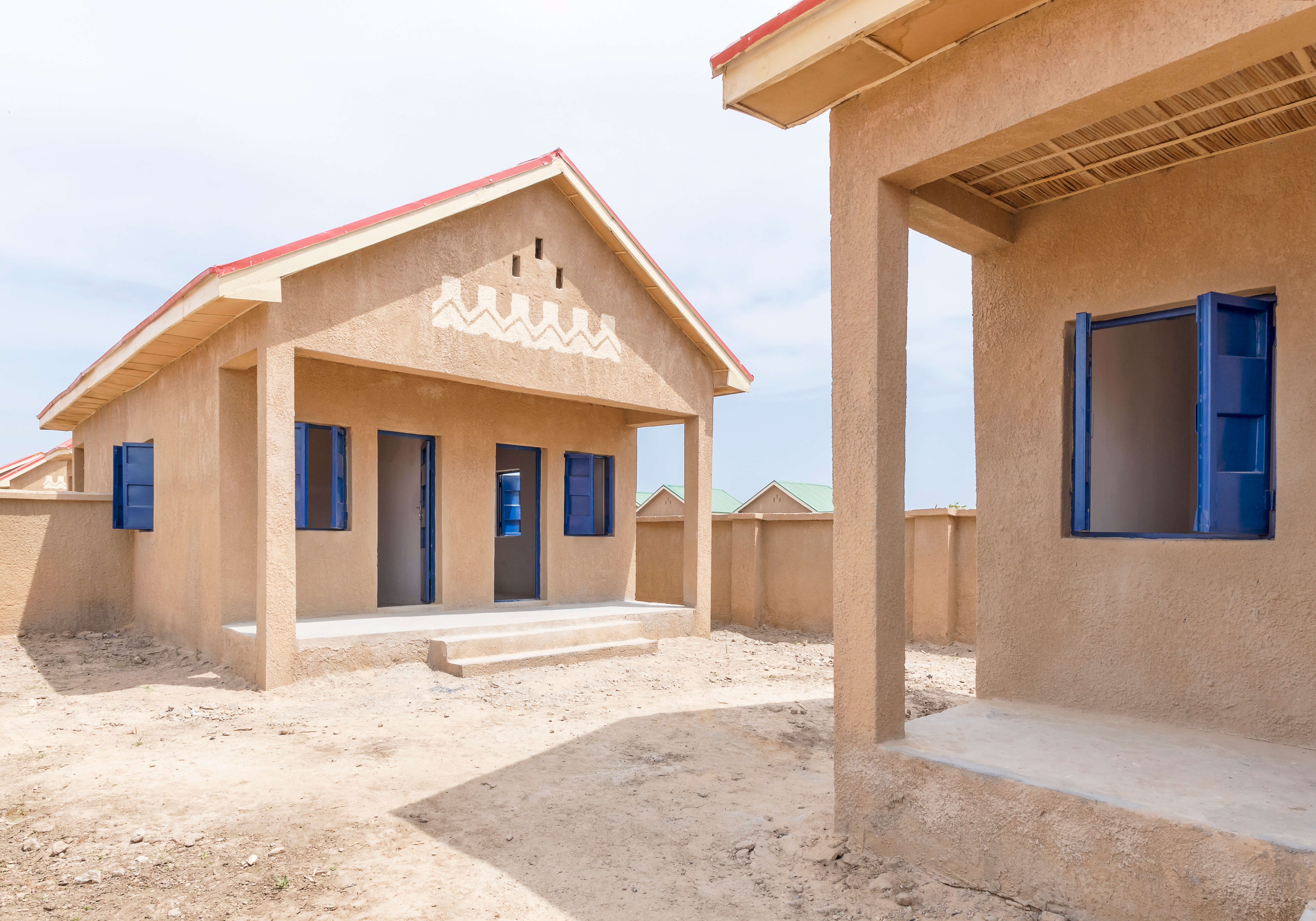 Architectural photograph of a housing project in Ngarannam, showcasing the design by Oshinowo Studio, a leading architectural design studio in Lagos, led by Tosin Oshinowo. The image features a row of new homes with sandy ochre finishes and distinct red roofs. The façades are decorated with traditional white geometric patterns above vibrant blue doors and matching window shutters, standing out against the clear sky. The structures are modern yet reflect local architectural styles, set within a dry landscape.