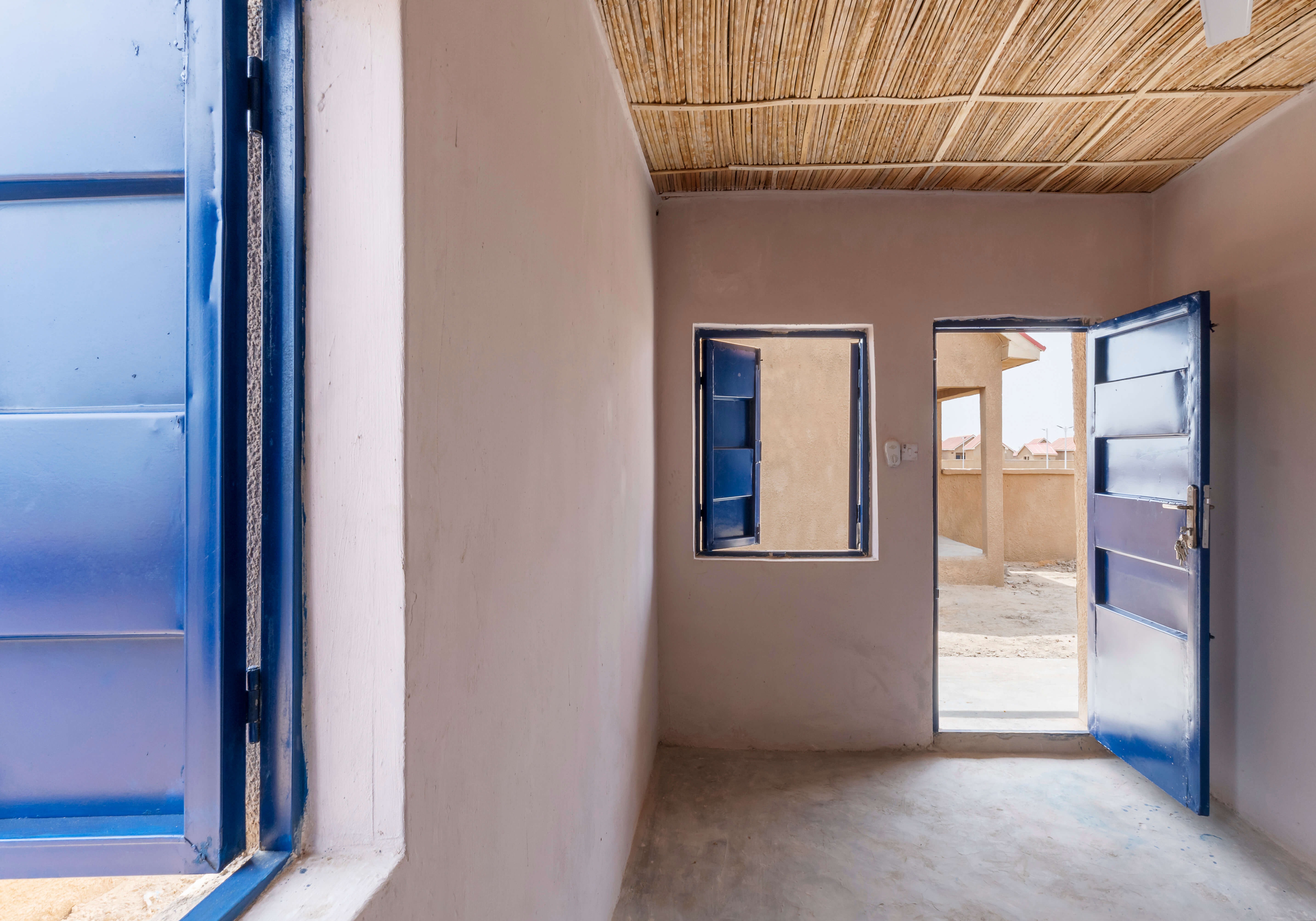 Interior view of a room designed by Oshinowo Studio, a leading architectural design studio in Lagos, led by Tosin Oshinowo, displaying a blend of traditional and contemporary elements. The room features a smooth concrete floor, earth-toned walls, and a natural thatched roof that adds texture and character to the space. A bright blue door stands open, coordinating with the color of the window shutters, inviting views of the adjacent rooms and the sandy exterior environment beyond.